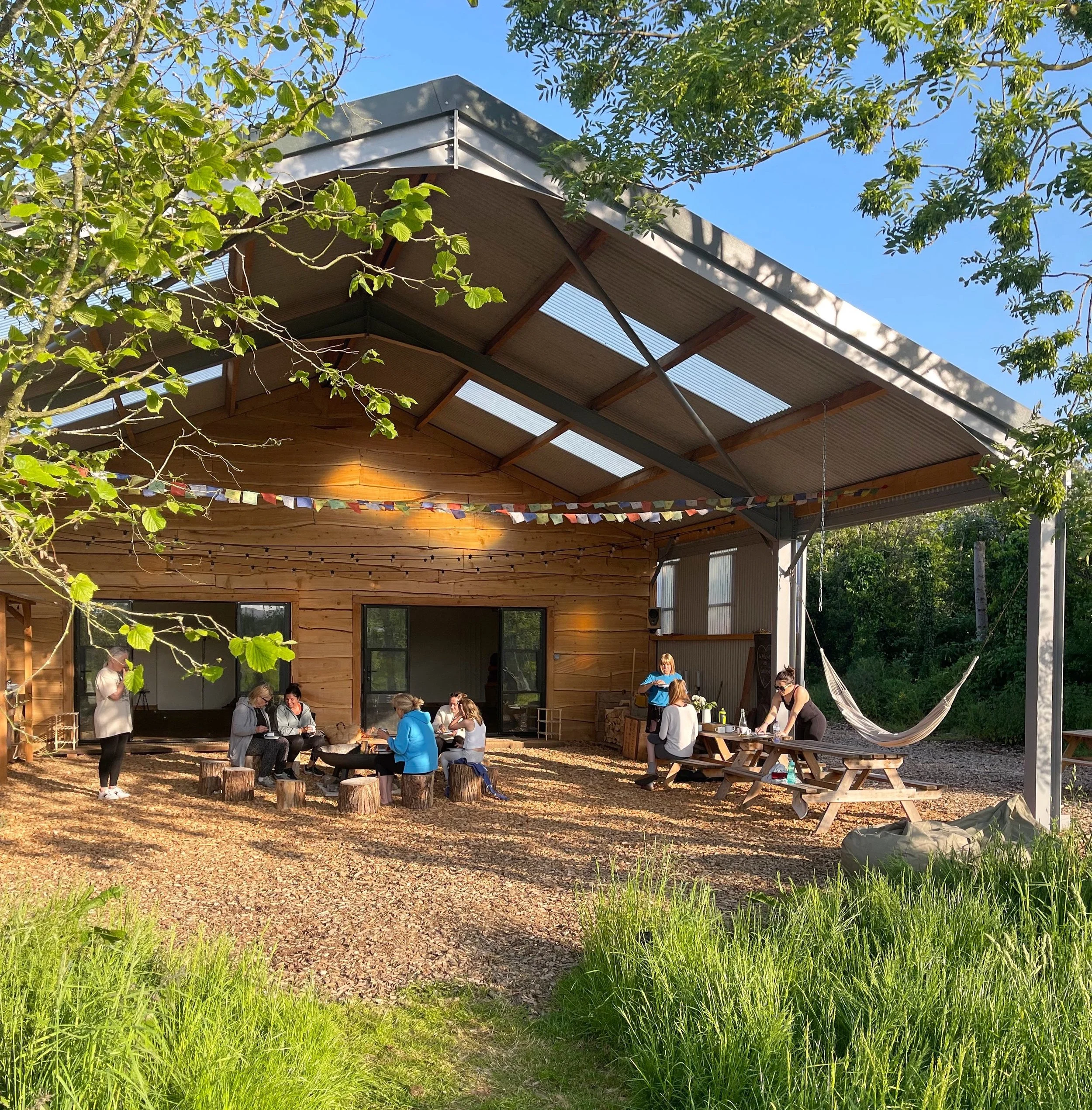 People gathered outdoors at a rustic wooden shelter with string lights, picnic tables, and a hammock, enjoying a social event on a sunny day surrounded by greenery.