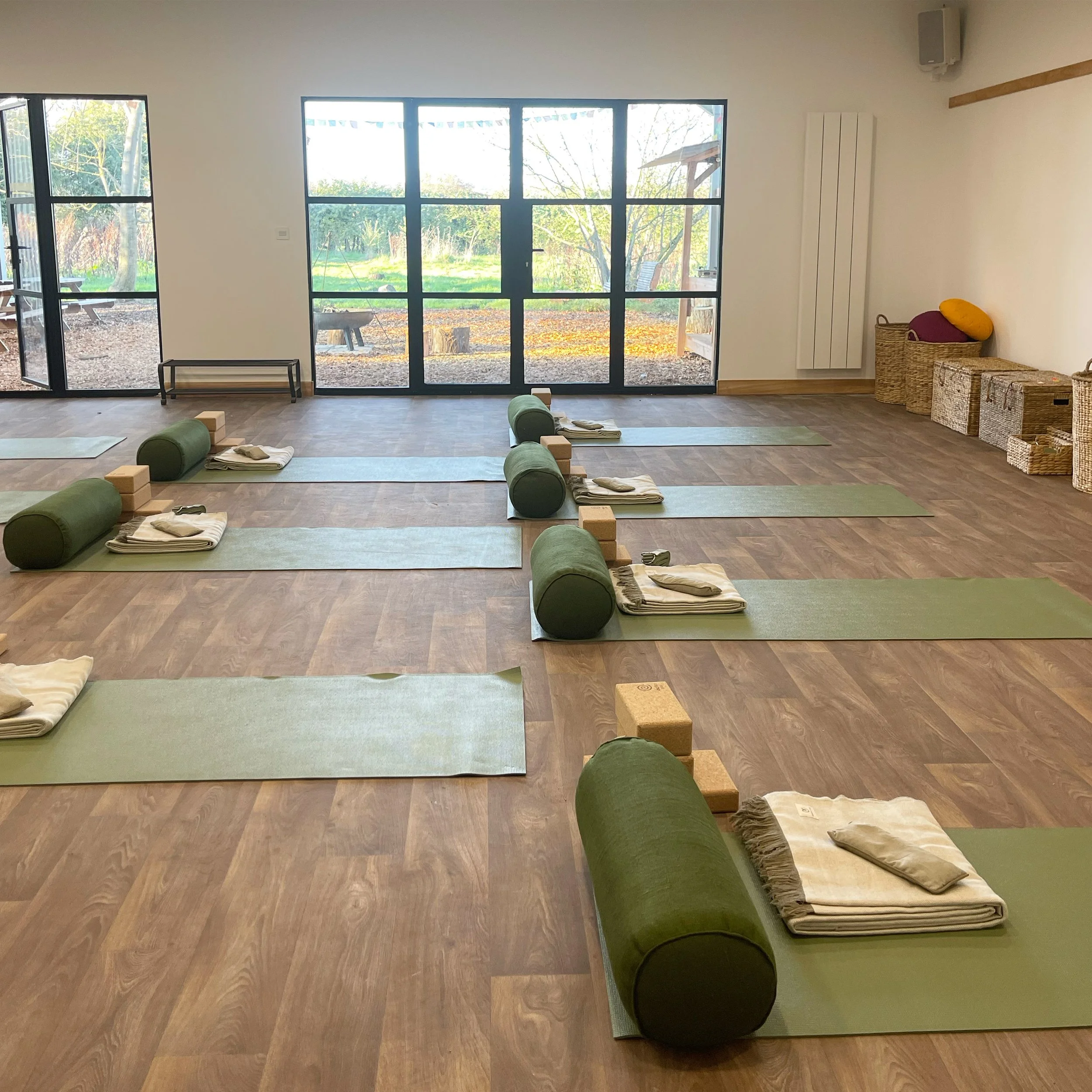 Yoga studio with yoga mats, bolsters, blocks, and blankets arranged on a wooden floor, with large windows showing an outdoor garden.