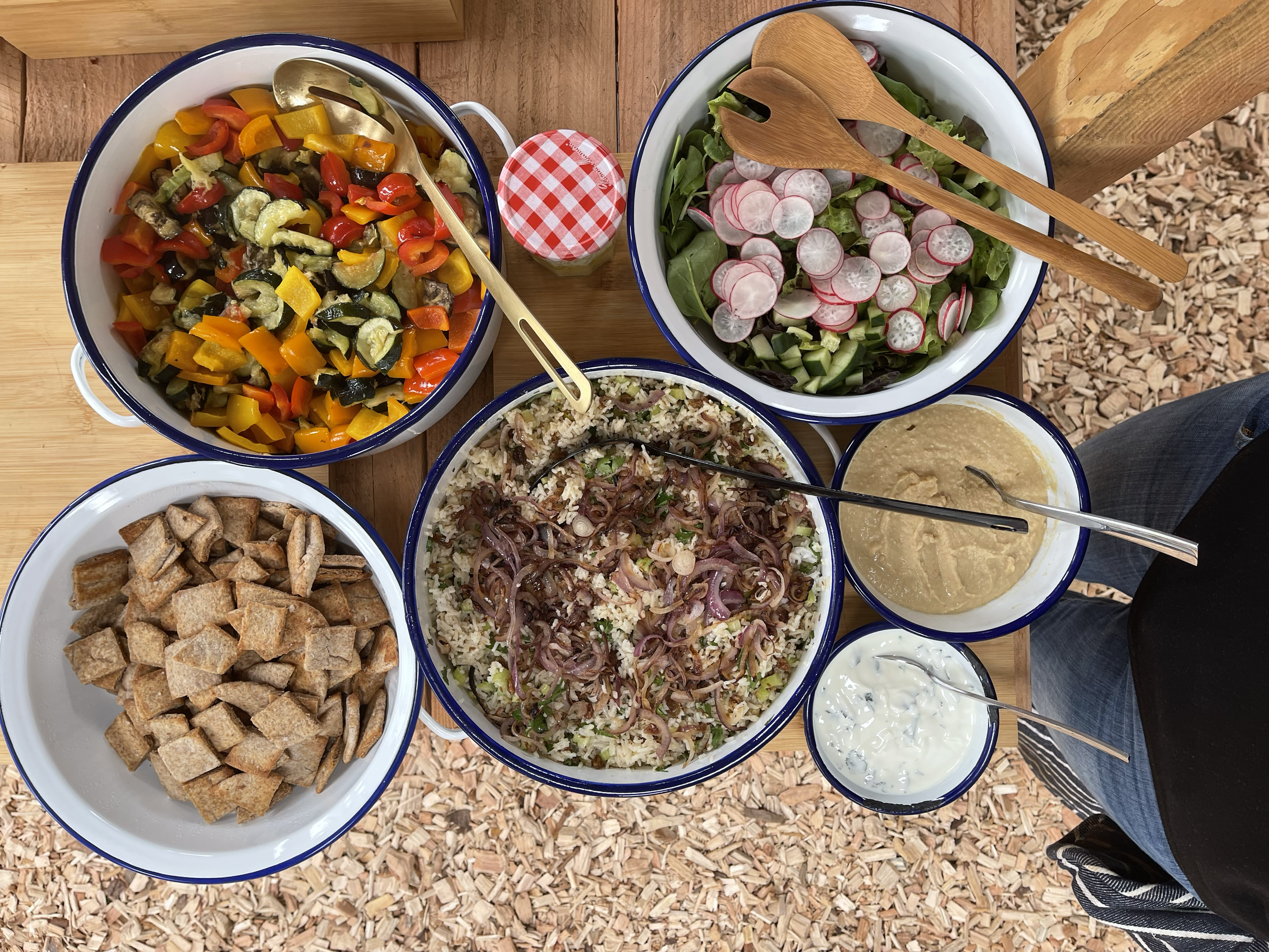 Top-down view of a table with various bowls of salad and toppings, including chopped vegetables, radishes, cucumbers, a rice dish topped with red onions, croutons, and condiments, on a wooden surface.