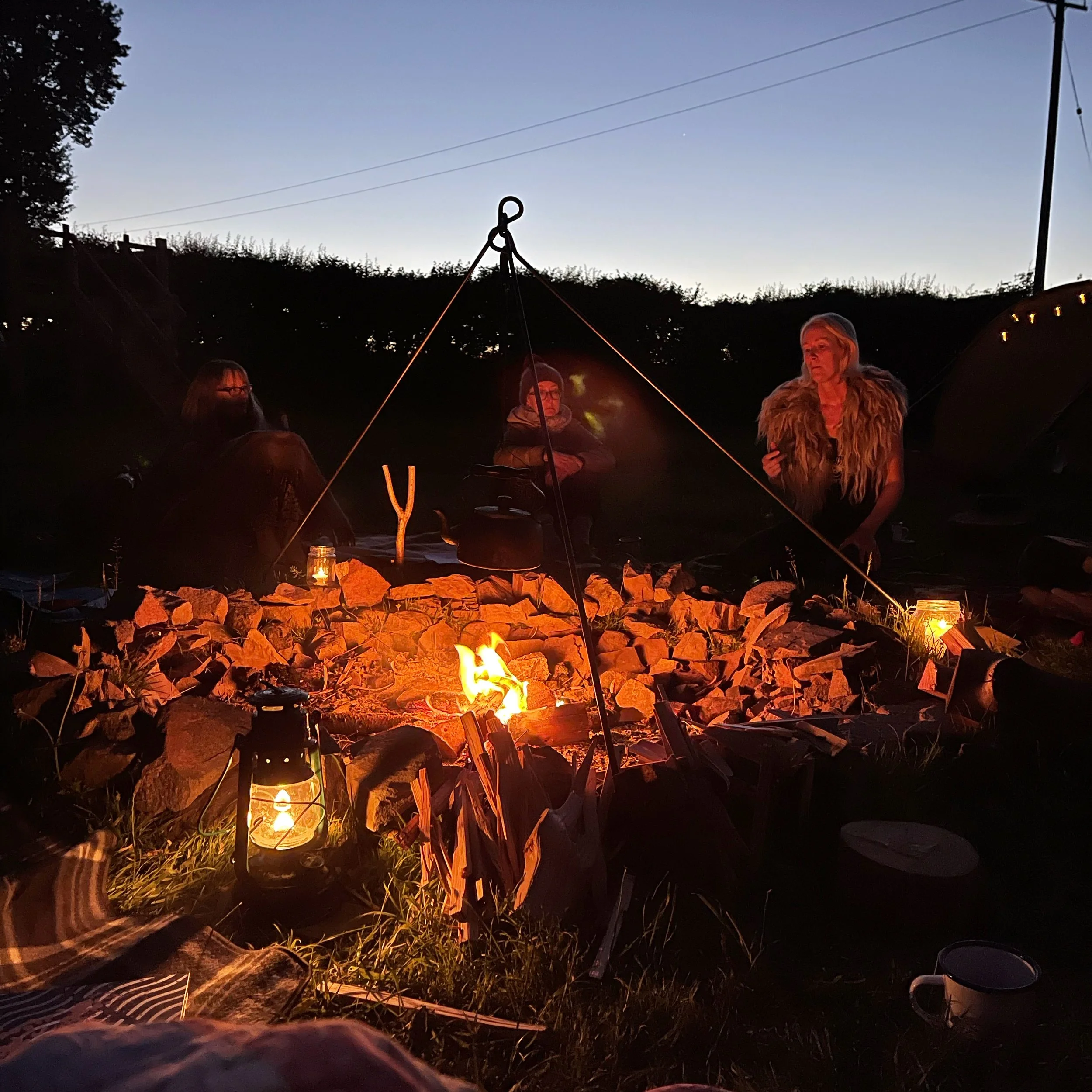Three women sitting around a campfire at dusk, with lanterns and a mug in the foreground.