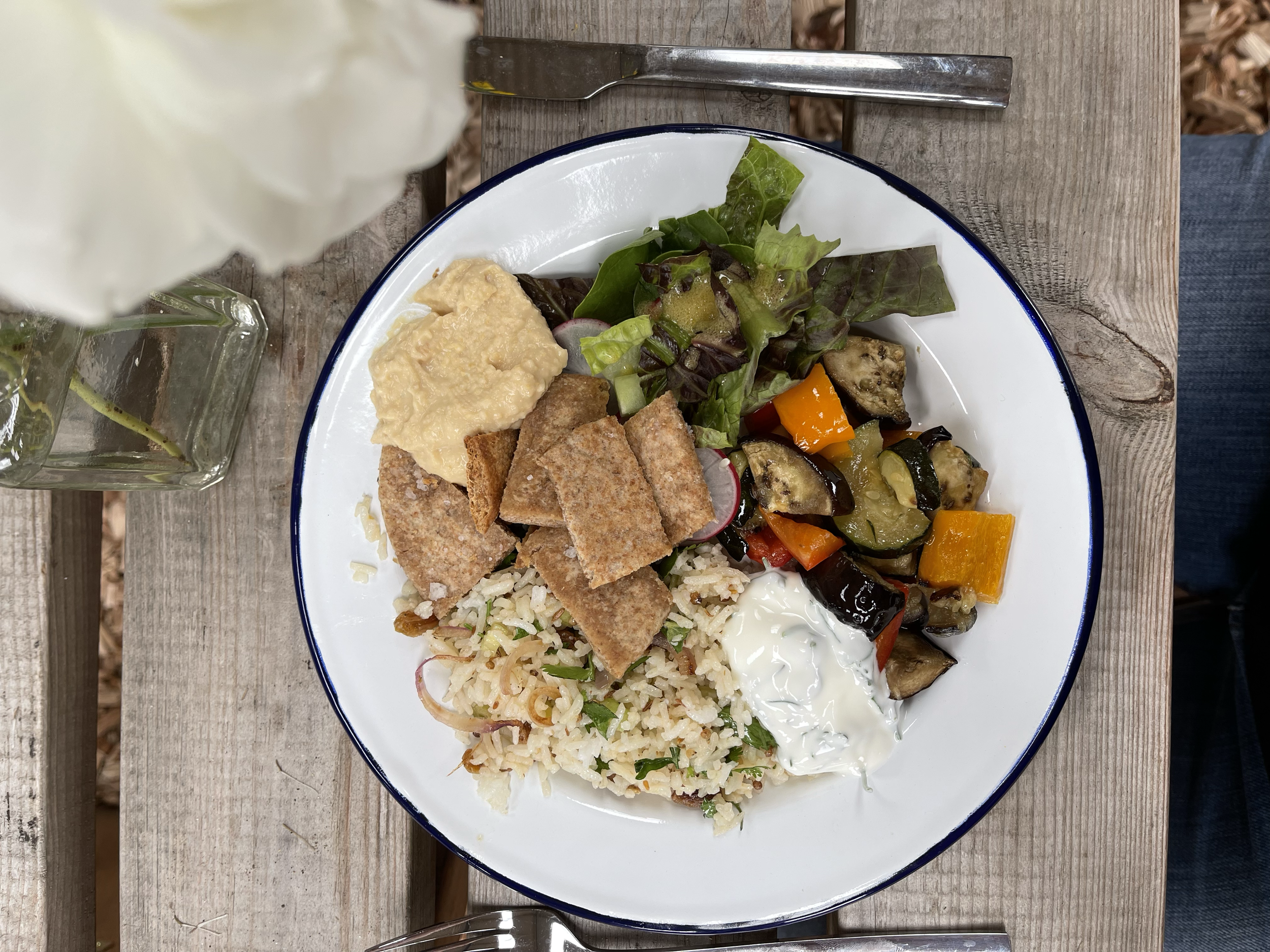 A plate of food with rice, a salad, roasted vegetables, hummus, and pita chips on a wooden table.