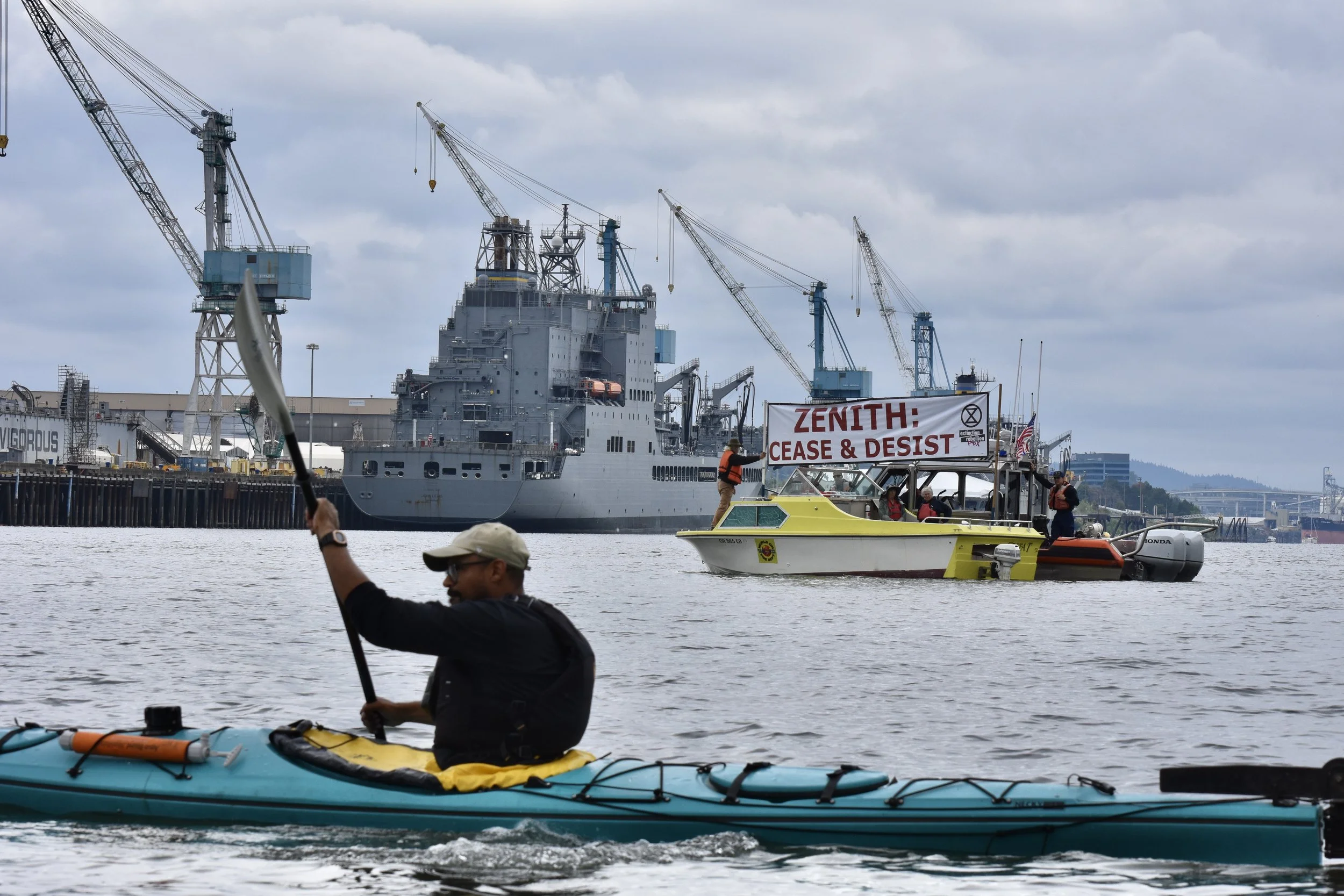 A paddler in a kayak in the foreground; in the background is a boat with a banner raised that says "Zenith: Cease and Desist!" Behind that, industrial boats and infrastructure.