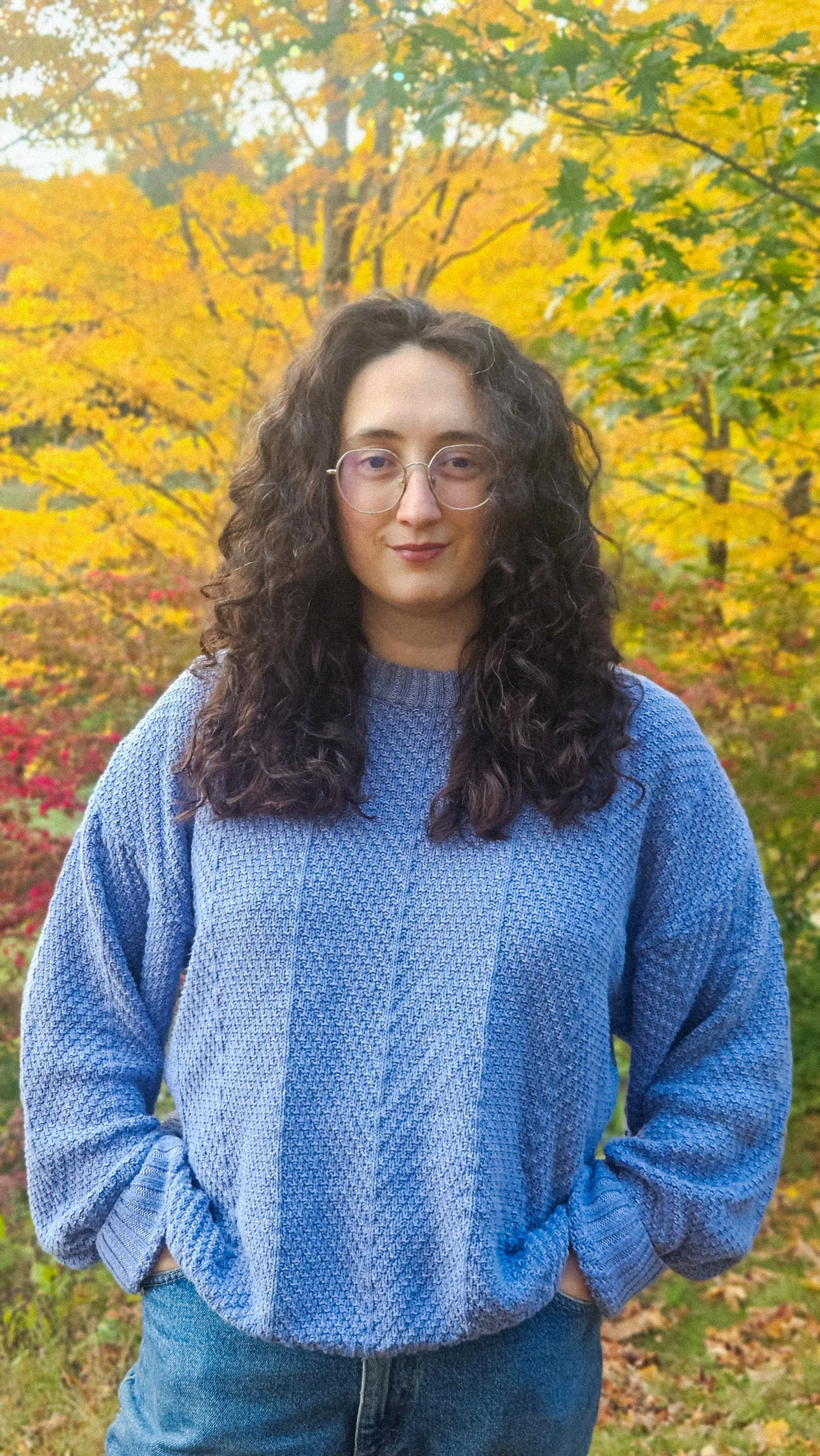 A white woman with glasses and curly brown hair, wearing a bright blue sweater, smiles gently amongst vibrant autumn foliage.