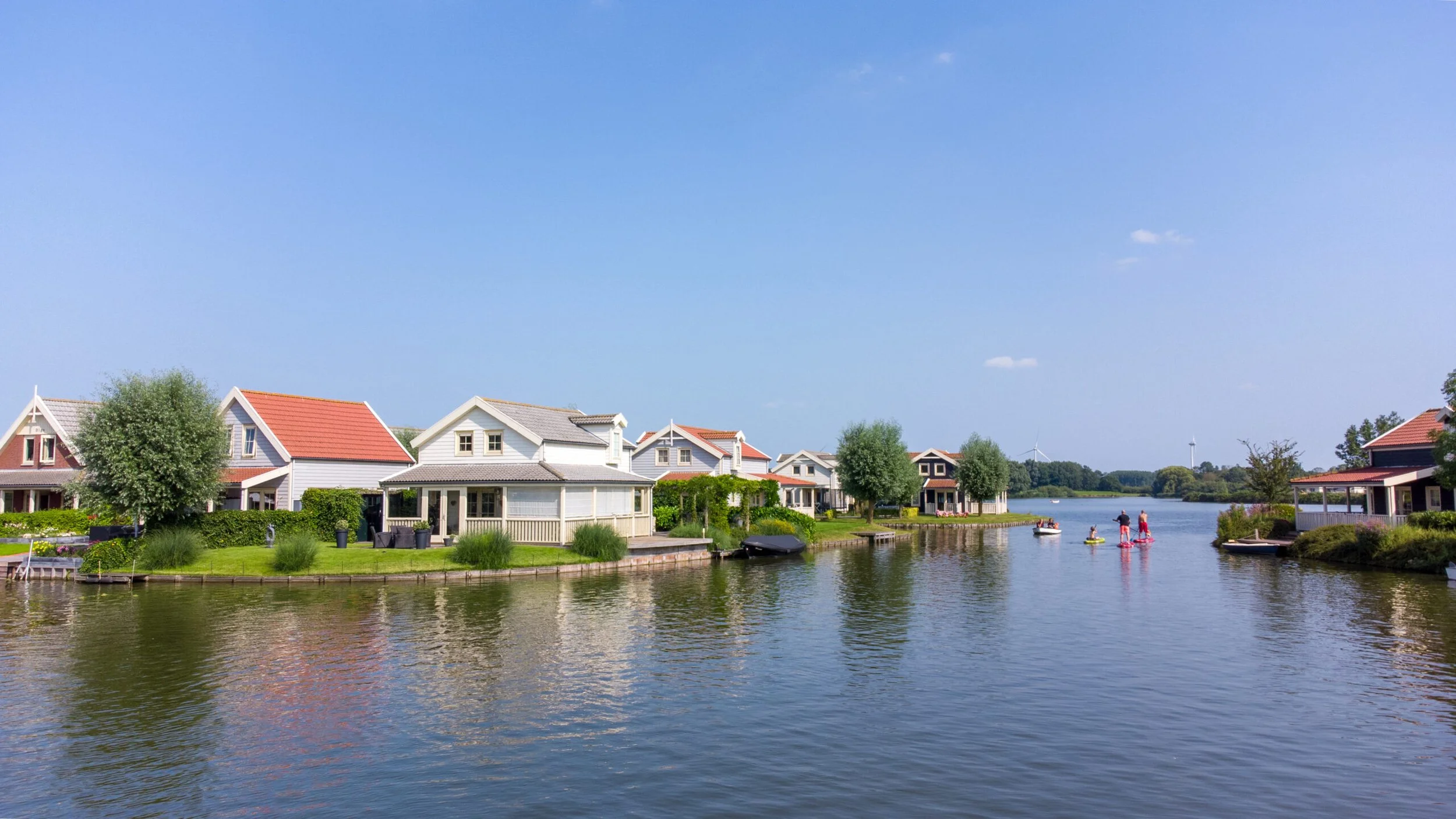 Ein uriges Dorf an einem Fluss mit weißen und bunten Häusern, grünen Bäumen und Menschen, die unter klarem blauen Himmel auf dem Wasser Boot fahren und paddeln.