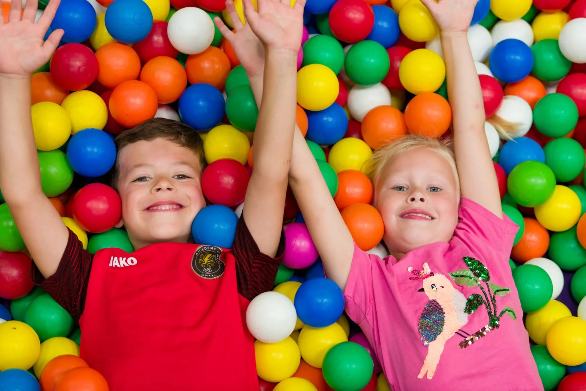 Two children, a boy and a girl, lie in a ball pit full of colorful plastic balls, laughing and holding hands.