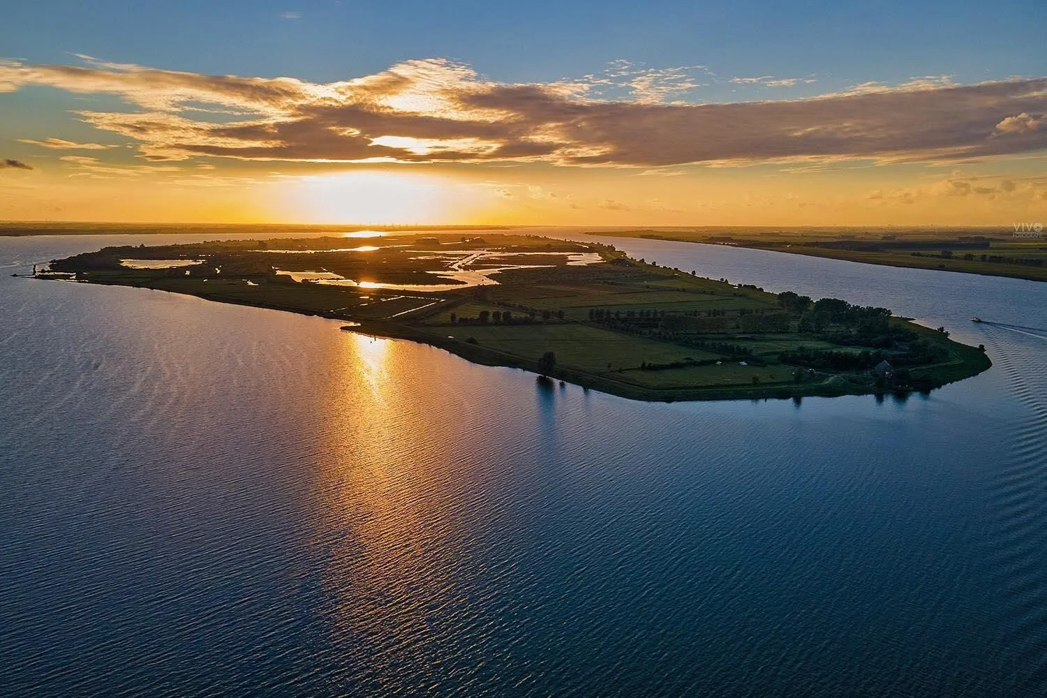 Landschaft mit Wasser und Land, Sonnenuntergang mit Wolken und Spiegelung auf dem Wasser, grüne Felder und schmale Kanäle in einer inselartigen Lage.