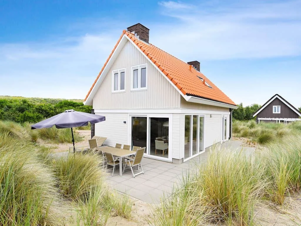 Ein weißes Haus mit rotem Dach, gelegen an einem Sandstrand mit umliegendem Gras, unter blauem Himmel.