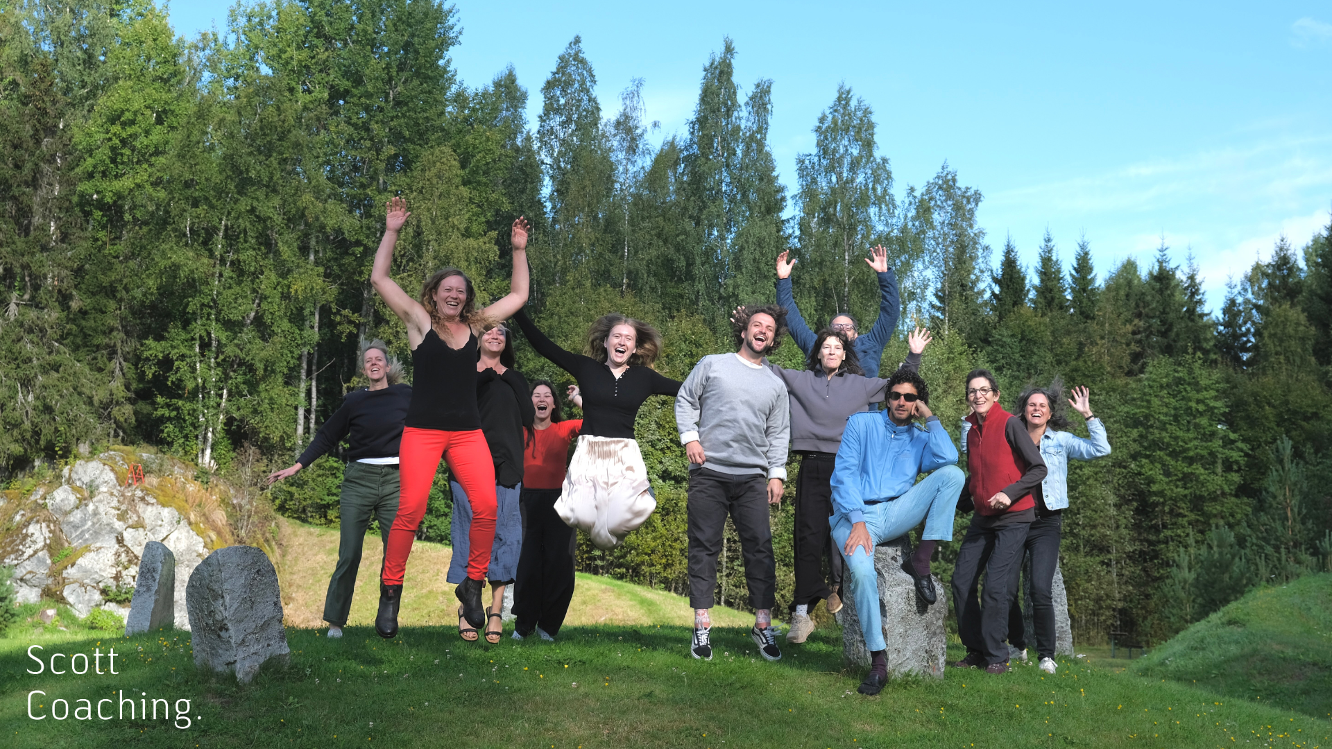 A group of artists at Arteles Creative Centre in Finland jumping and laughing together outdoors, surrounded by green trees and blue sky, capturing a joyful residency moment.