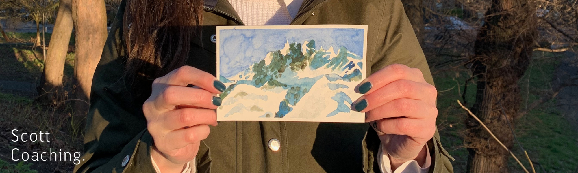 Close-up photo of hands with teal-colored nail polish holding up a small watercolor painting of mountains. The painting features snow-capped peaks in white and blue-green tones against a light blue sky.