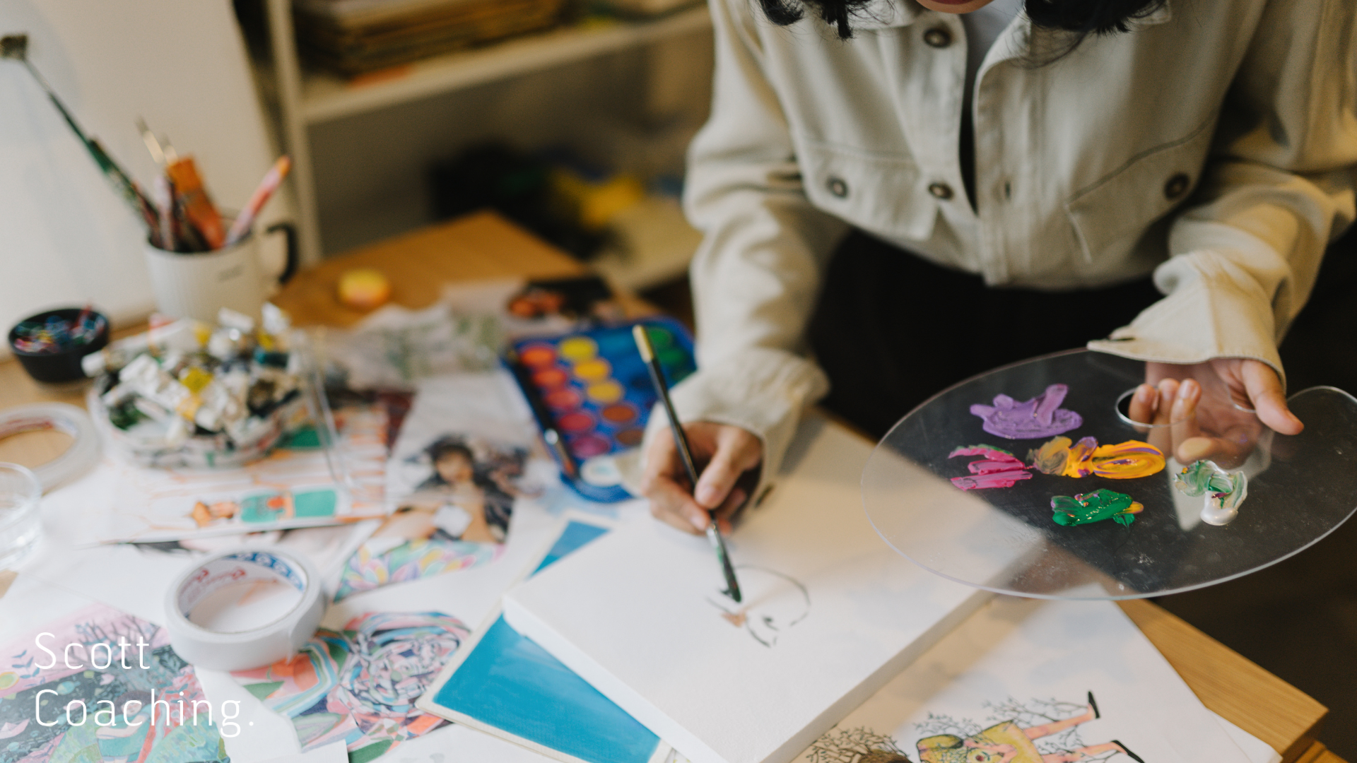 A person painting at a cluttered desk covered in art supplies, including paint tubes, brushes, watercolours, and illustrated papers, with a clear palette of vibrant paint colours in one hand and a brush in the other.