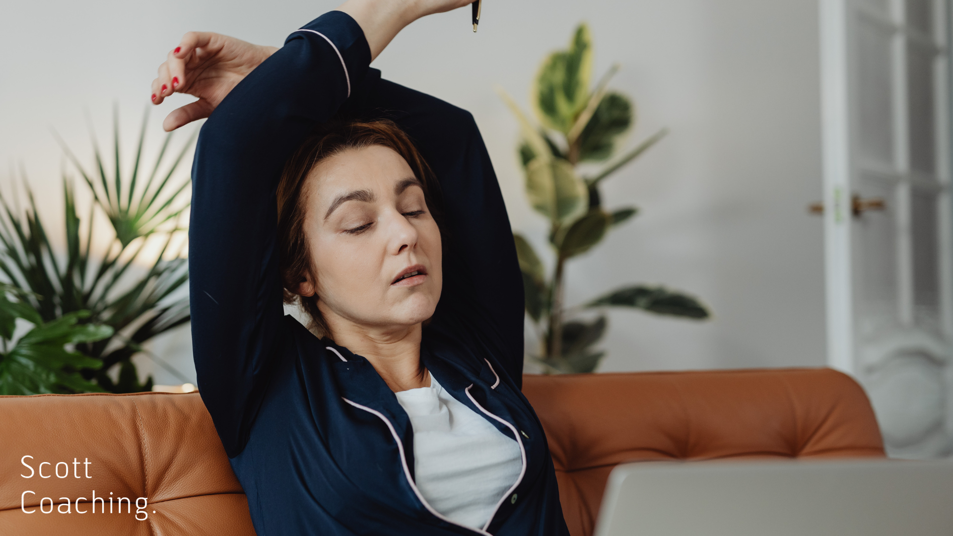Woman in navy loungewear stretching on an orange couch with eyes closed, plants in background, embodying rest and self-care