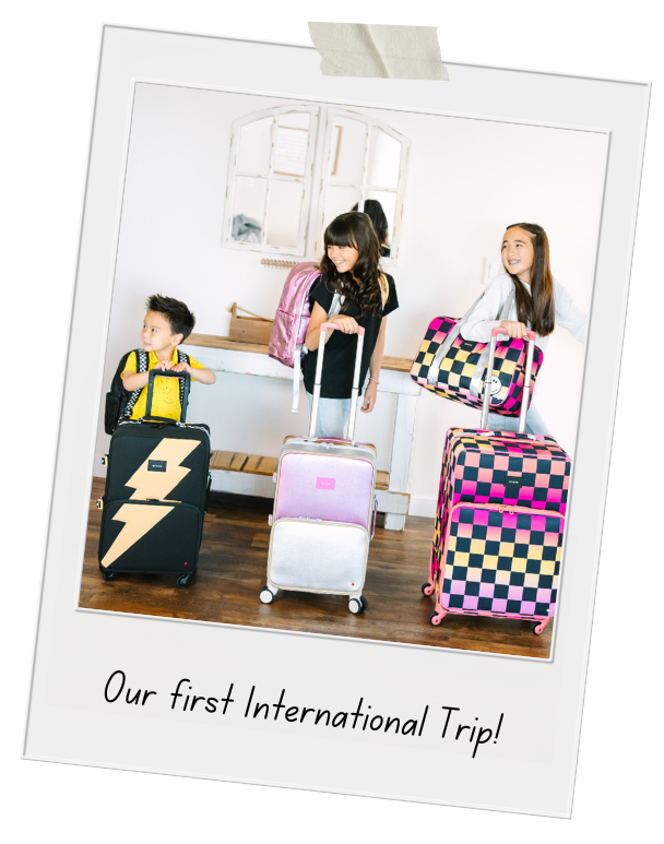 Three children with suitcases and backpacks standing indoors by a large window, smiling and excited, celebrating their first international trip.