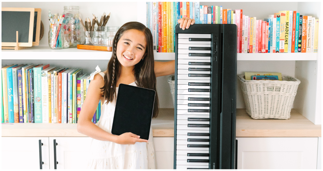 A young girl with long dark hair holding a black tablet, smiling, standing in front of a large keyboard/workstation in a room with bookshelves filled with colorful books and jars.