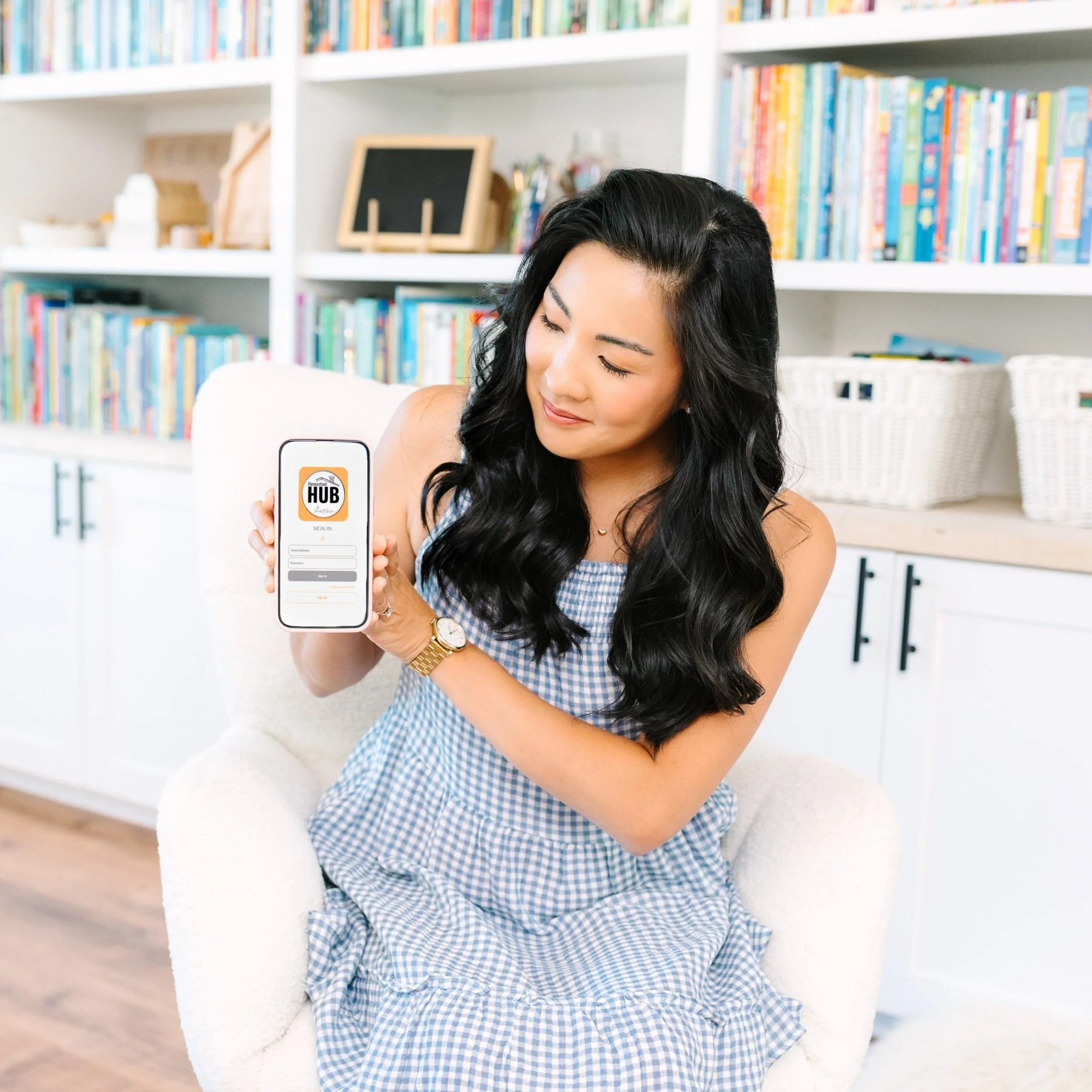 A woman with long black wavy hair, wearing a blue and white checkered dress, is sitting on a white chair in front of a bookshelf filled with colorful books. She is holding a smartphone displaying an app called HUB, and she is smiling softly.