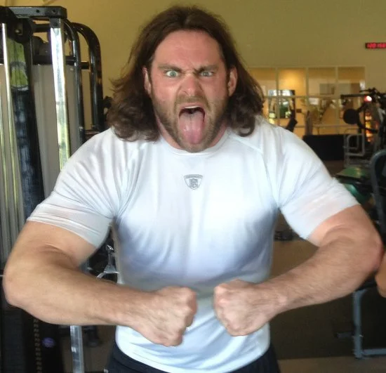 Man with long brown hair, wearing a white t-shirt, making a fierce face with tongue out and fists clenched in gym.