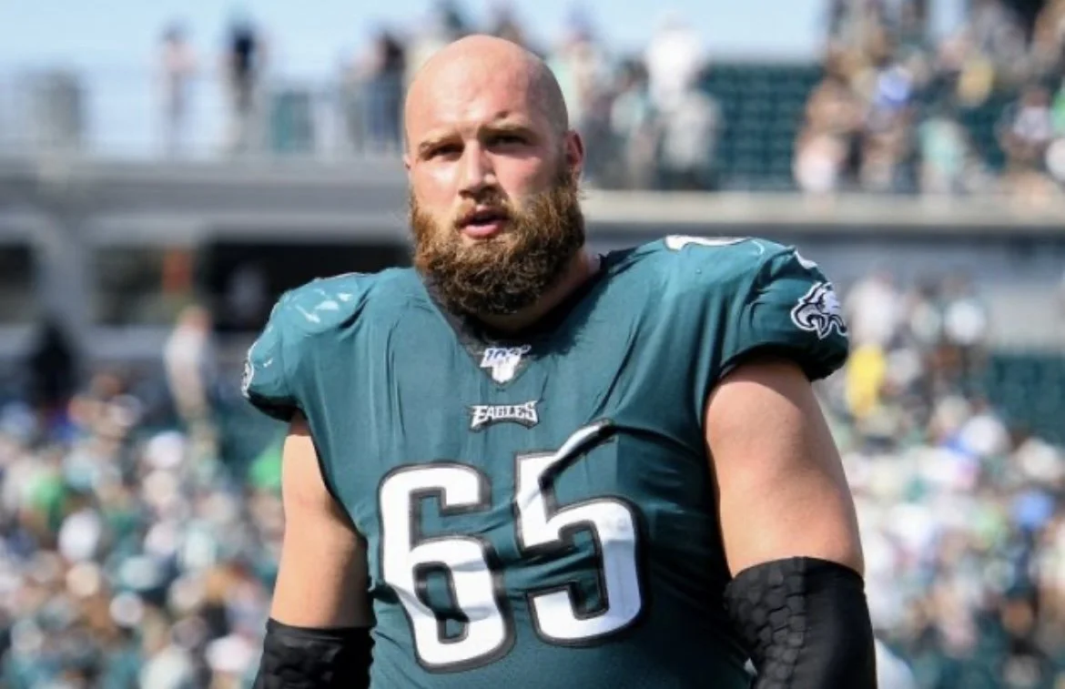 A football player in a teal jersey with the number 65 and a beard, on the field with a blurred crowd in the background.