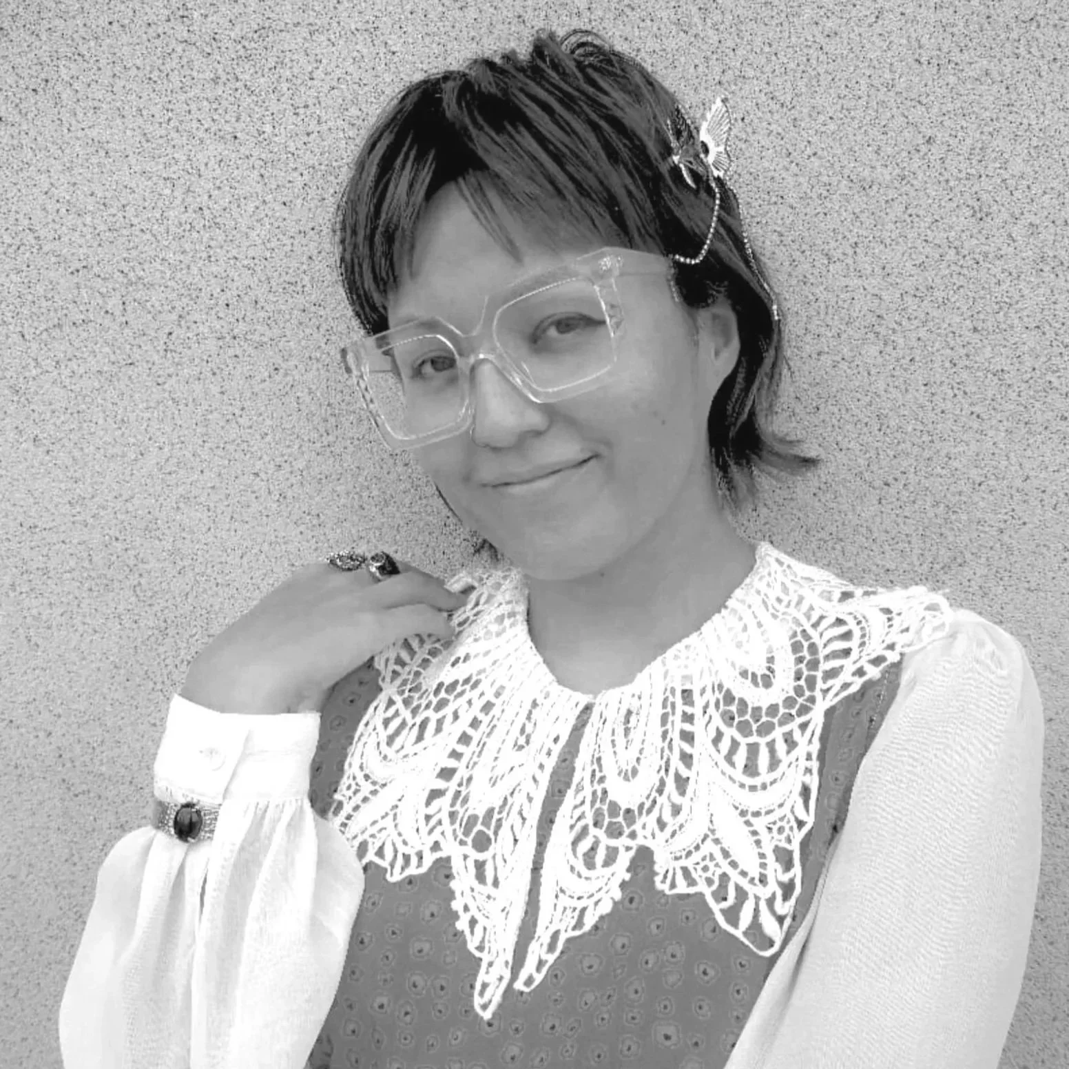 A woman with short dark hair wearing large glasses, vintage earrings, and a lace-collared blouse, smiling and posing against a textured wall.