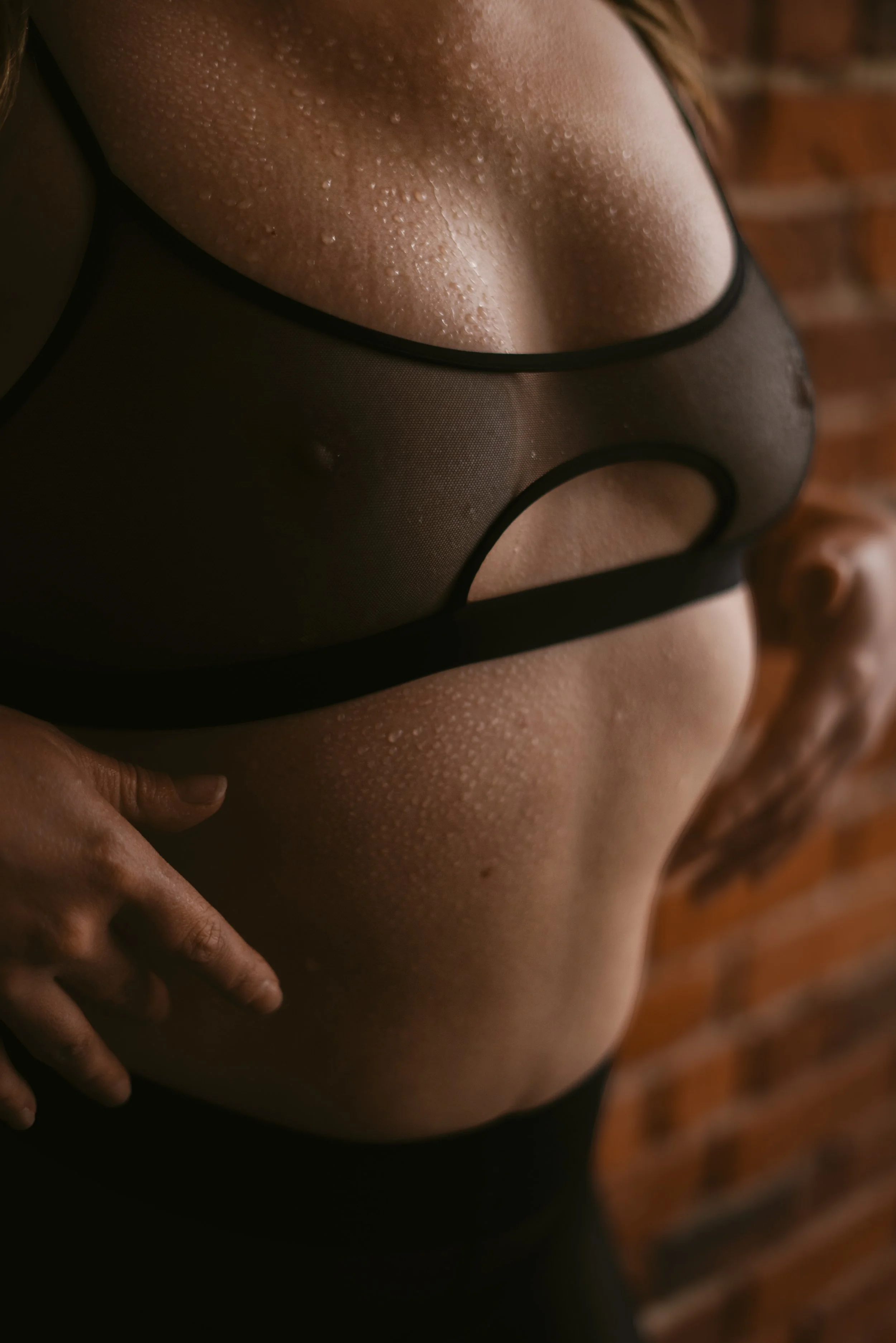 Close-up of a wet, sweaty torso of a person wearing a black, sheer, lingerie top with a pierced nipple, against a blurred brick wall background.