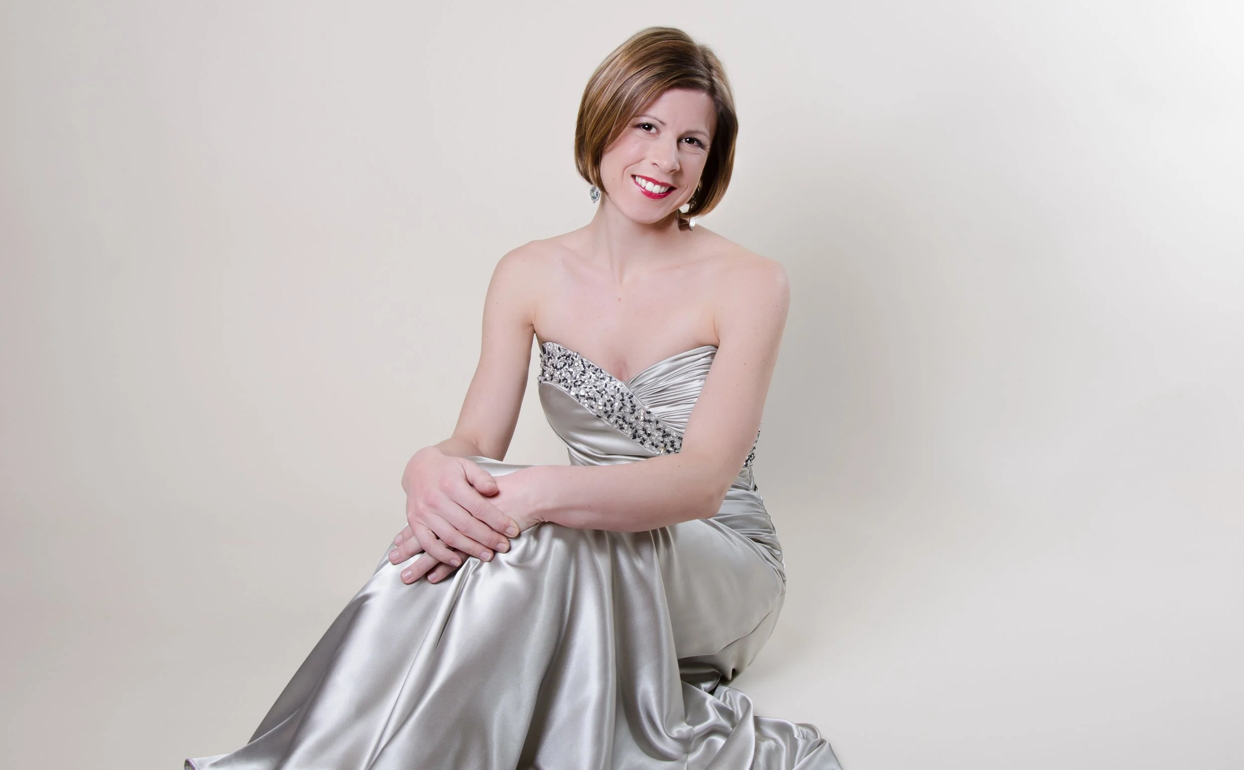 A woman in a strapless, silver satin gown with rhinestone embellishments on the bodice, sitting against a plain white background, smiling at the camera.