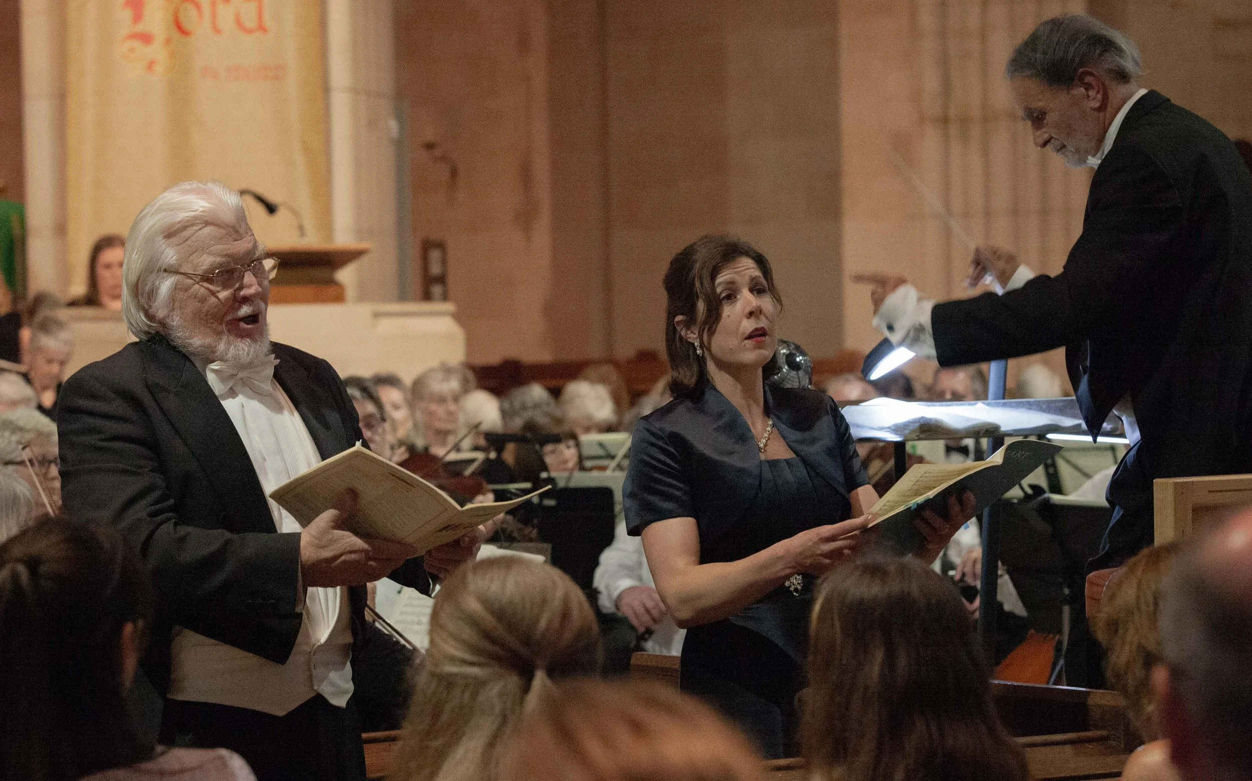 A choir performs in a concert hall with an orchestra in the background, three singers in the foreground with sheet music, conductor directing them.