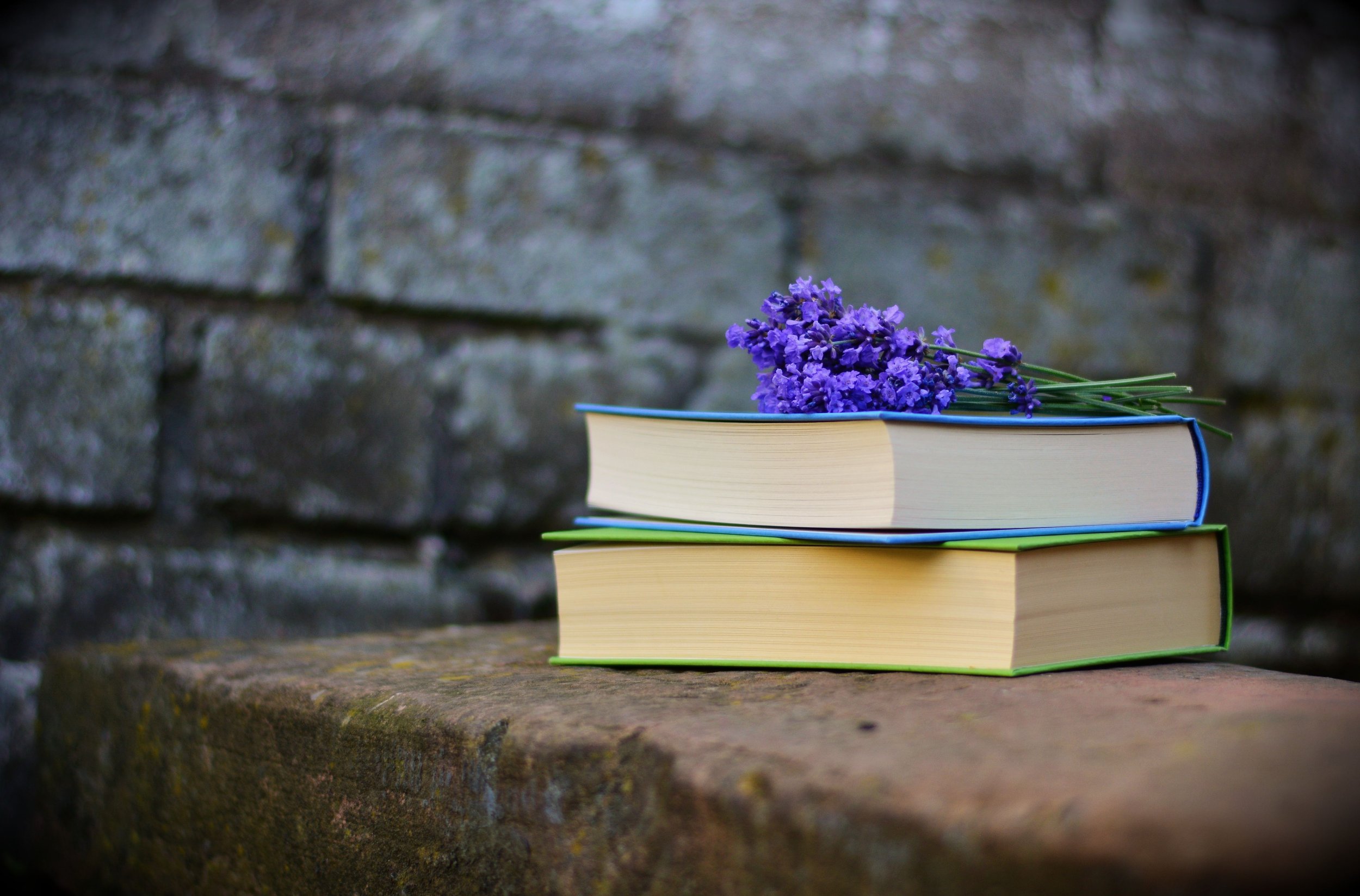 A stack of three books with a small bouquet of purple flowers on top, placed on a stone surface against a blurred stone wall background.