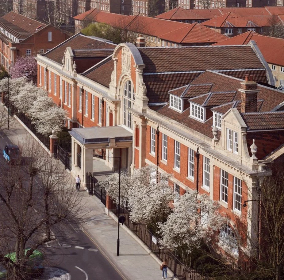 Aerial view of a red brick school or institutional building with a large arched window and decorative architecture, surrounded by trees with white blossoms, on a city street.