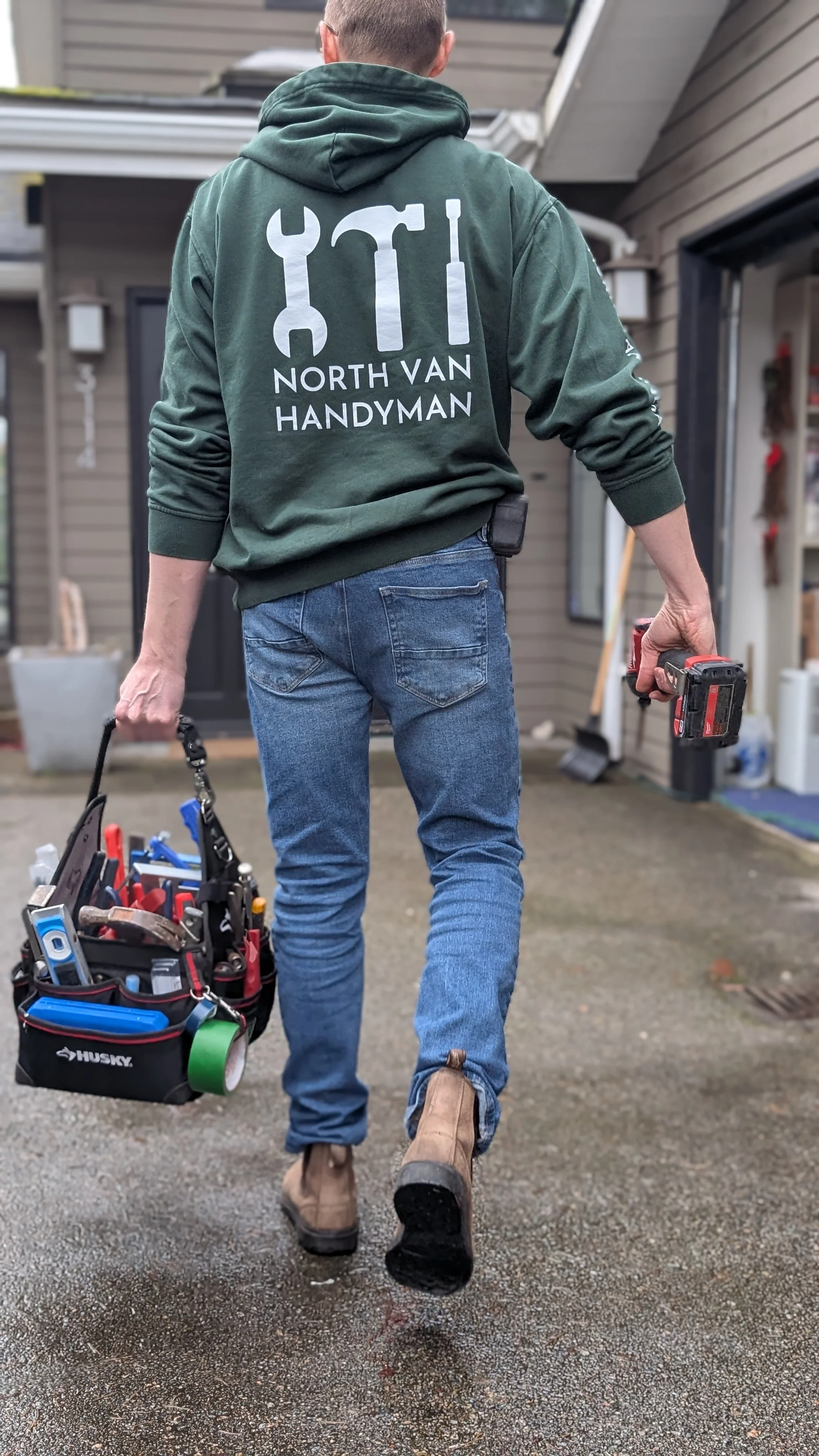 Back view of a handyman wearing a green hoodie with 'North Van Handyman' and tools logos on the back, carrying a tool bag and a cordless drill, walking on a wet concrete driveway in front of a house.