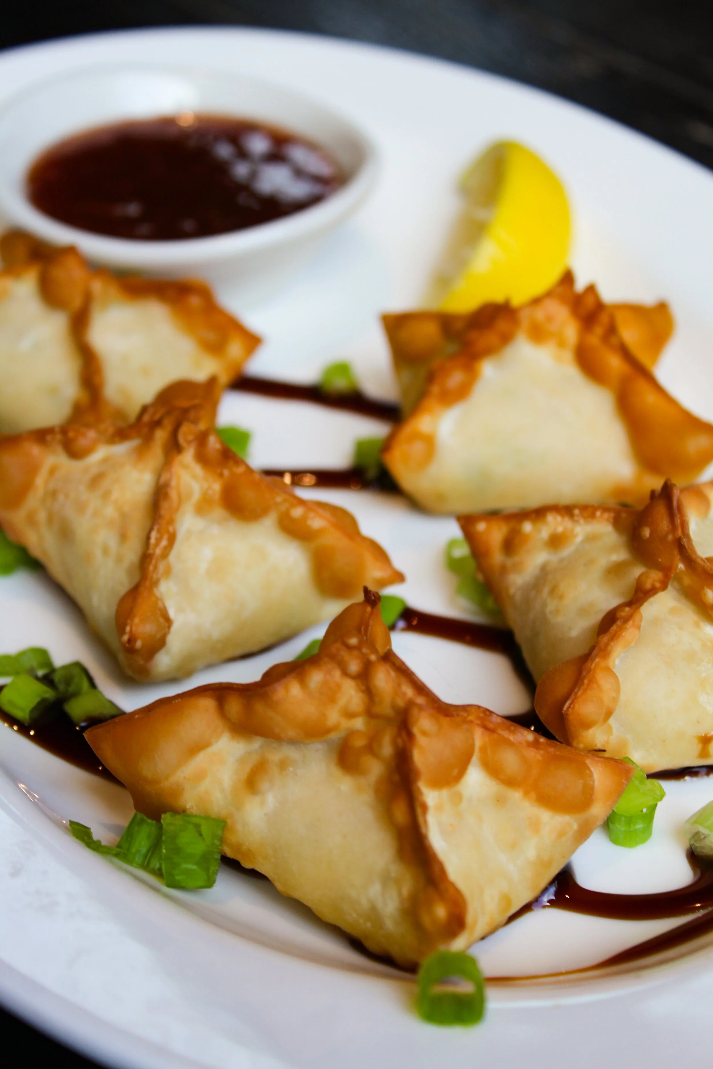 Close-up of five fried wontons arranged on a white plate, garnished with chopped green onions, accompanied by a small dish of dark dipping sauce and a lemon wedge in the background.