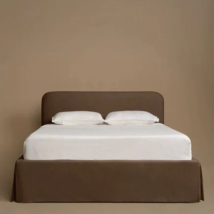Modern upholstered platform bed in warm taupe velvet with a softly rounded headboard, tailored bed skirt, and crisp white bedding against a neutral backdrop.