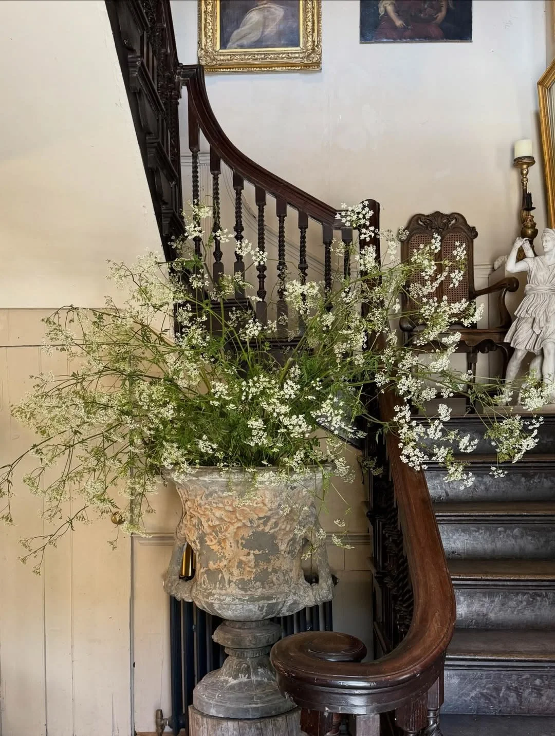 Traditional luxury entryway with a handcrafted timber staircase, antique stone urn overflowing with seasonal wildflowers, classical artwork, and vintage furnishings, celebrating timeless craftsmanship and natural beauty.
