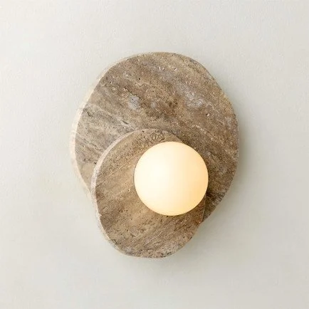 Sculptural natural stone wall sconce with layered organic forms and a glowing frosted glass globe, mounted on a warm white wall.