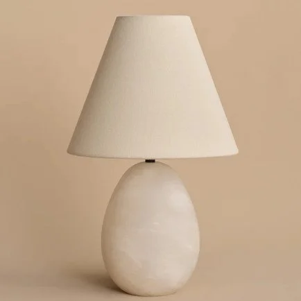 Sculptural alabaster table lamp with an oval stone base and a tapered cream fabric shade against a warm neutral background.