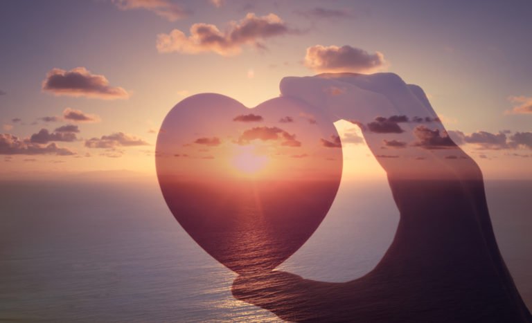 person holds heart shaped object overlaid on image of sunset over the ocean