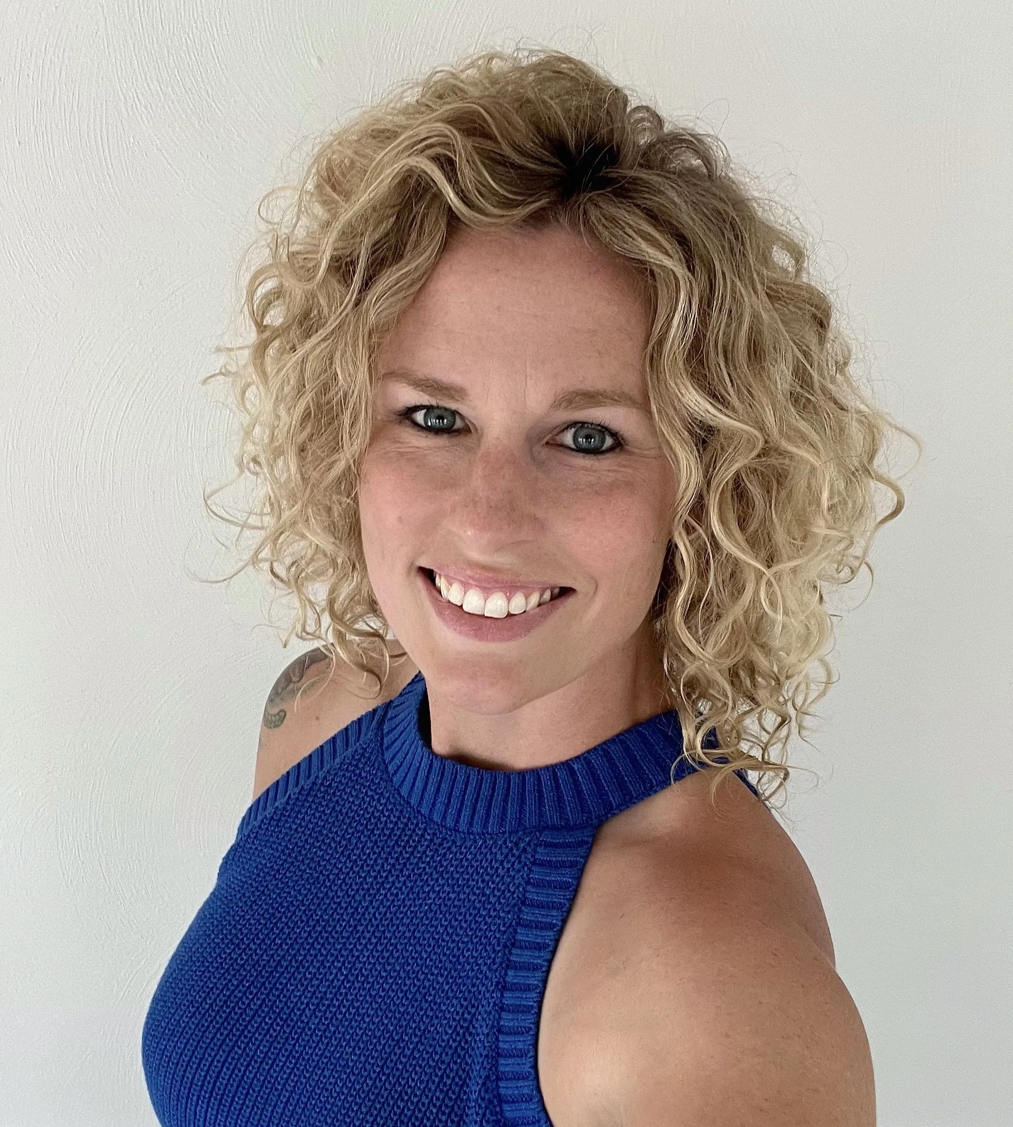 A woman with curly blonde hair, blue eyes, and a bright smile, wearing a sleeveless blue top, standing against a plain white wall.