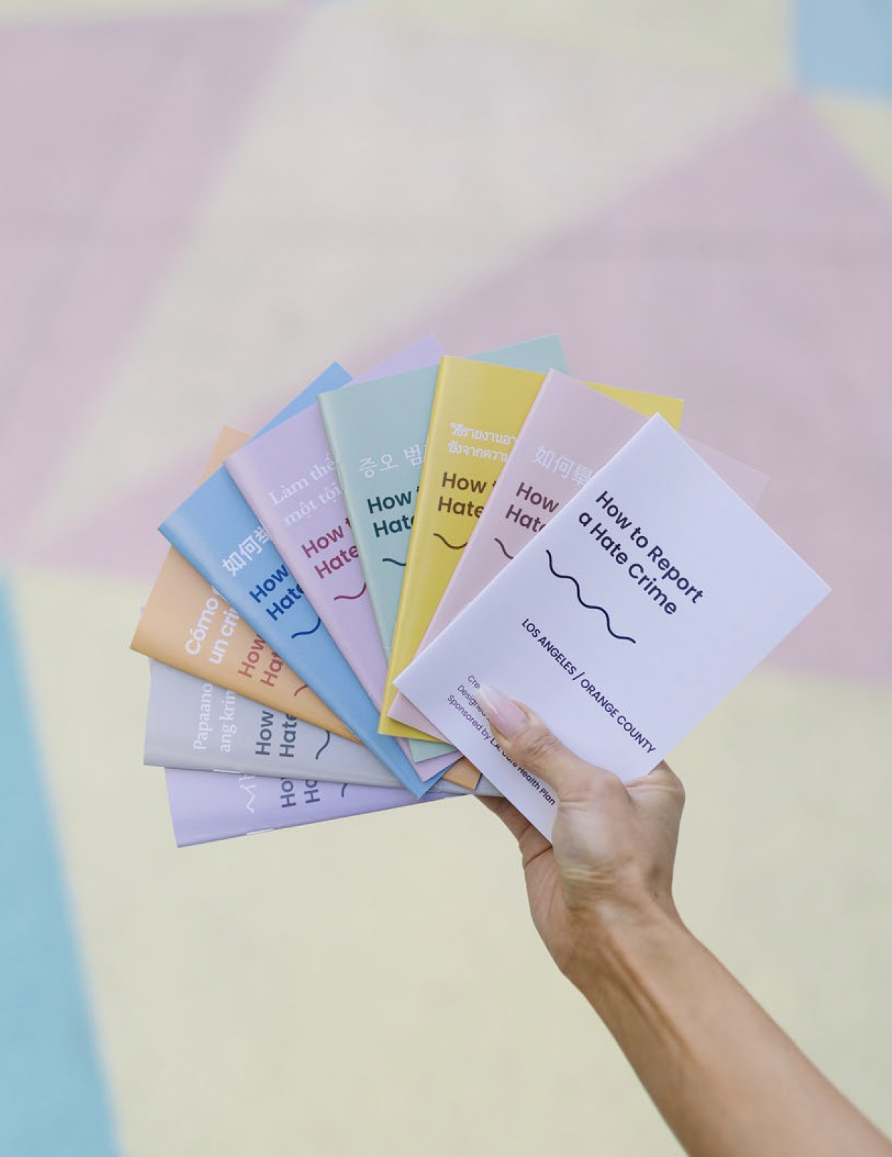 Hand holding a fan of colorful pamphlets titled 'How to Report a Hate Crime' with locations in Los Angeles and Orange County.
