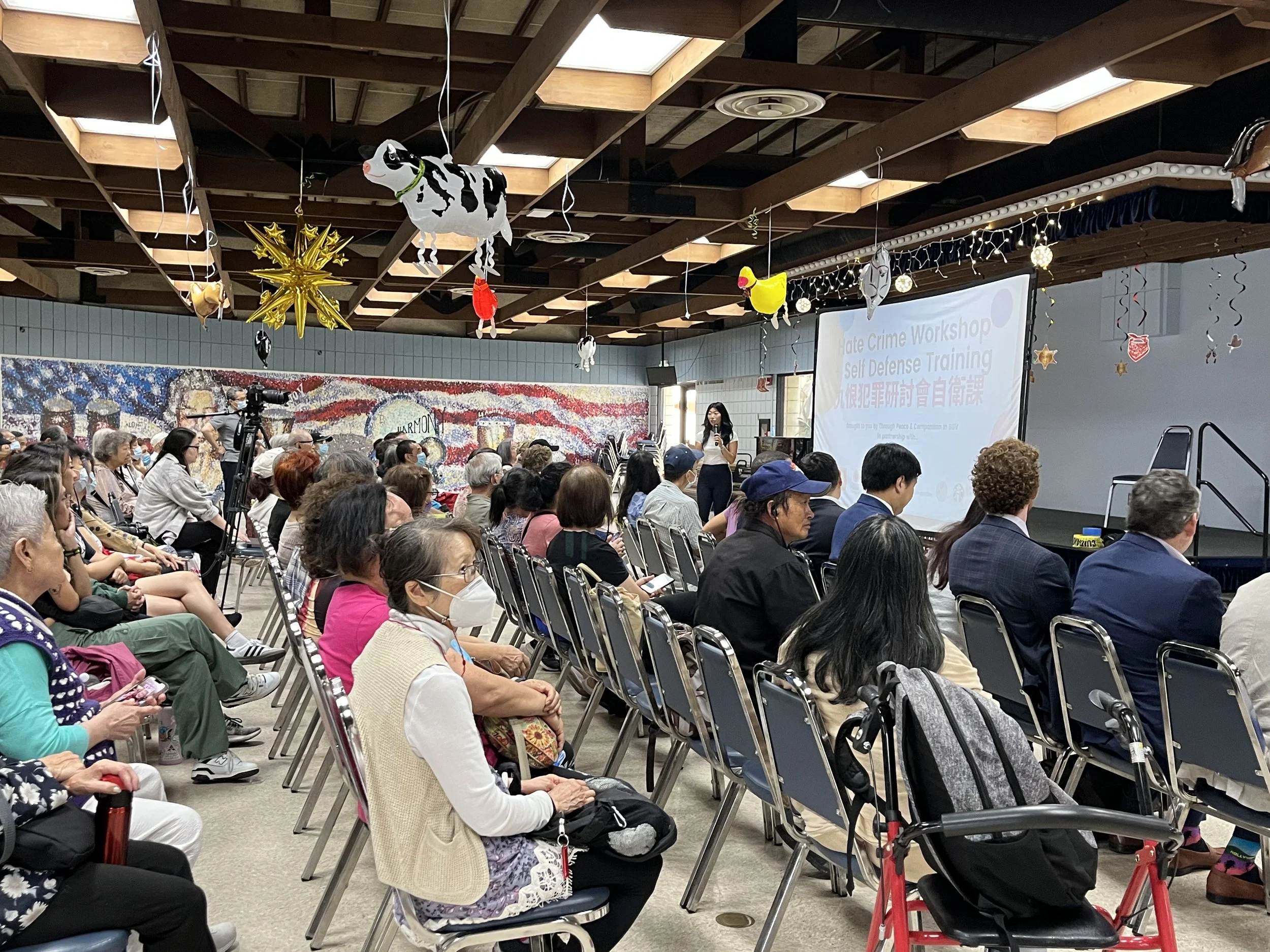 A large indoor gathering of diverse people, mostly seated and wearing masks, attending a Hate Crime Safety Workshop in a spacious room decorated with hanging animals and star-shaped balloons.