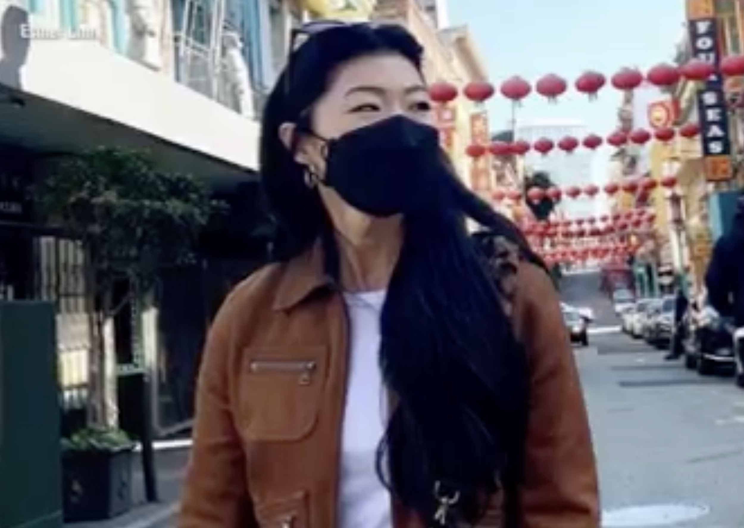Esther wearing a black face mask and brown jacket walking on a street decorated with red lanterns.