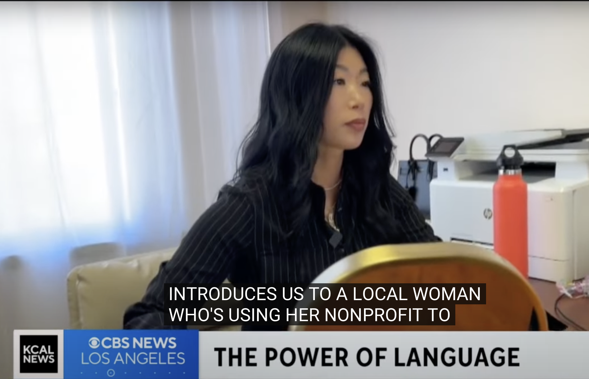 Esther is wearing a black striped shirt, sitting at a table in an office with printer and other equipment behind her, being interviewed by CBS News in Los Angeles about her nonprofit.