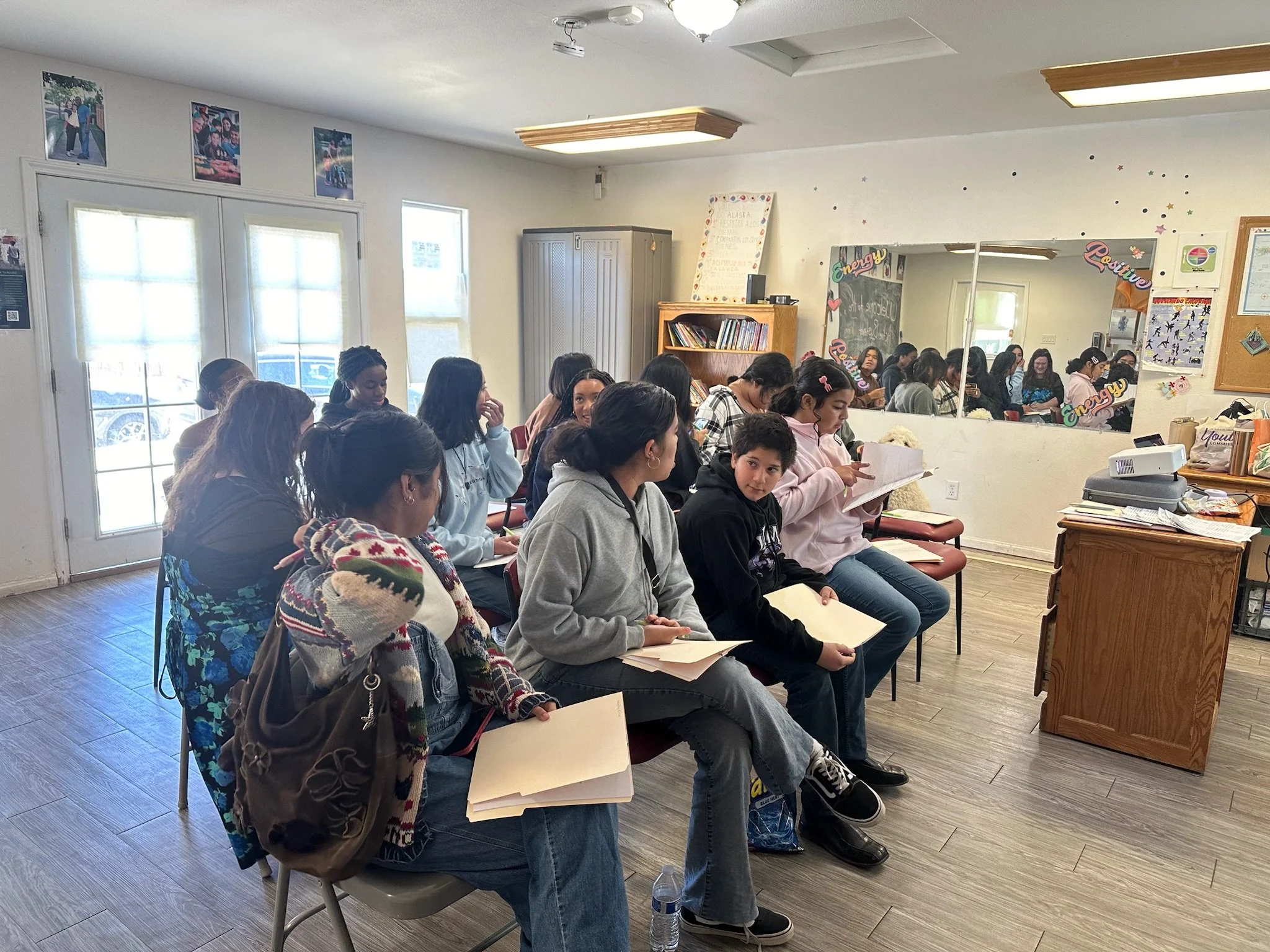 A group of students seated in a classroom, for an Active Bystander Youth Training Workshop. The classroom has white walls, a wooden door with glass panes, and a shelf with books.