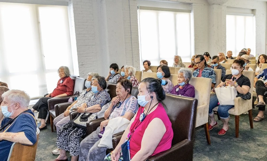 Group of elderly and middle-aged people sitting in an auditorium-like room, all wearing face masks, attending an Elder Abuse and Fraud Workshop.