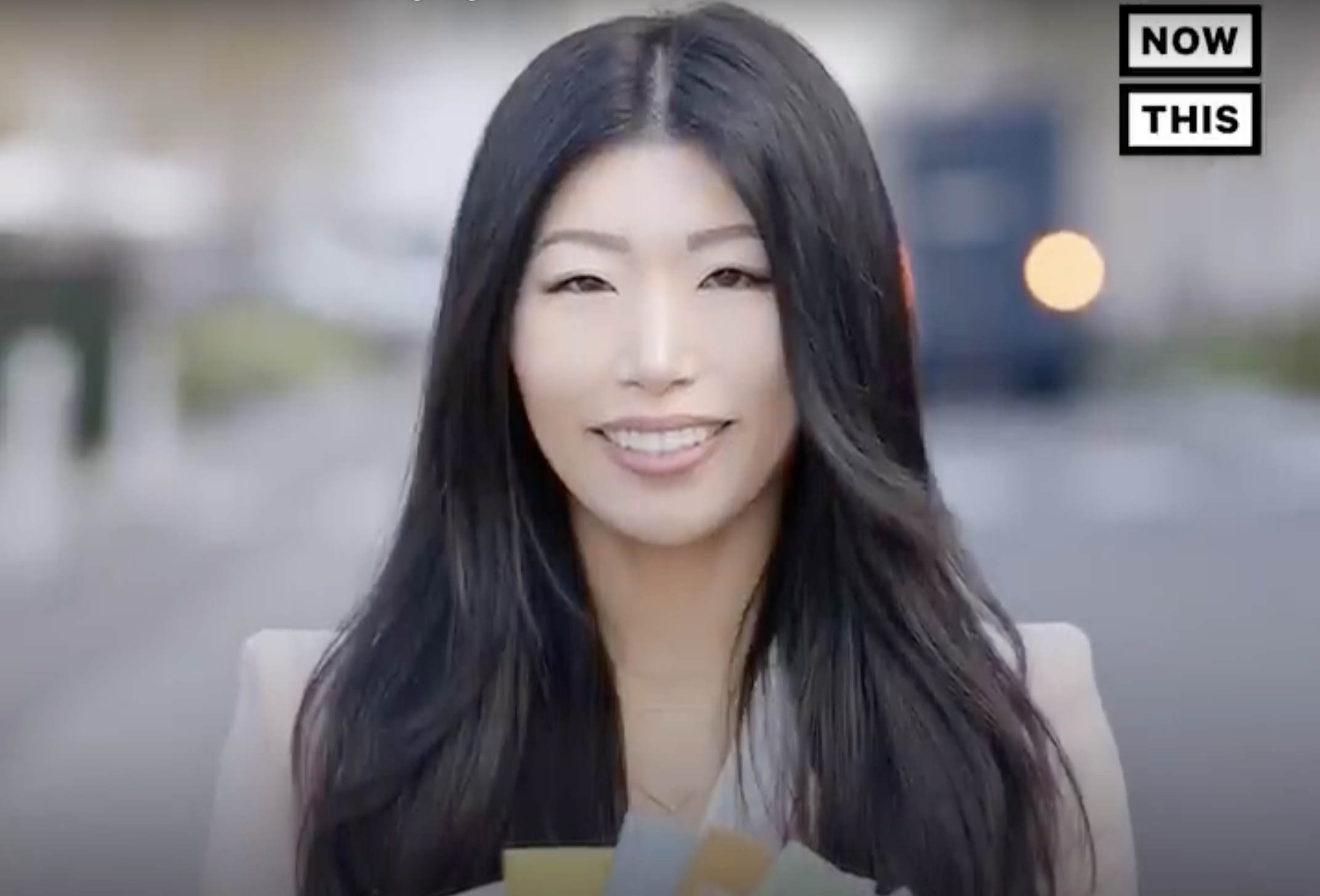 Esther with long black hair standing outdoors, with blurred background and an overlay graphic that reads "NOW THIS" in black and white.