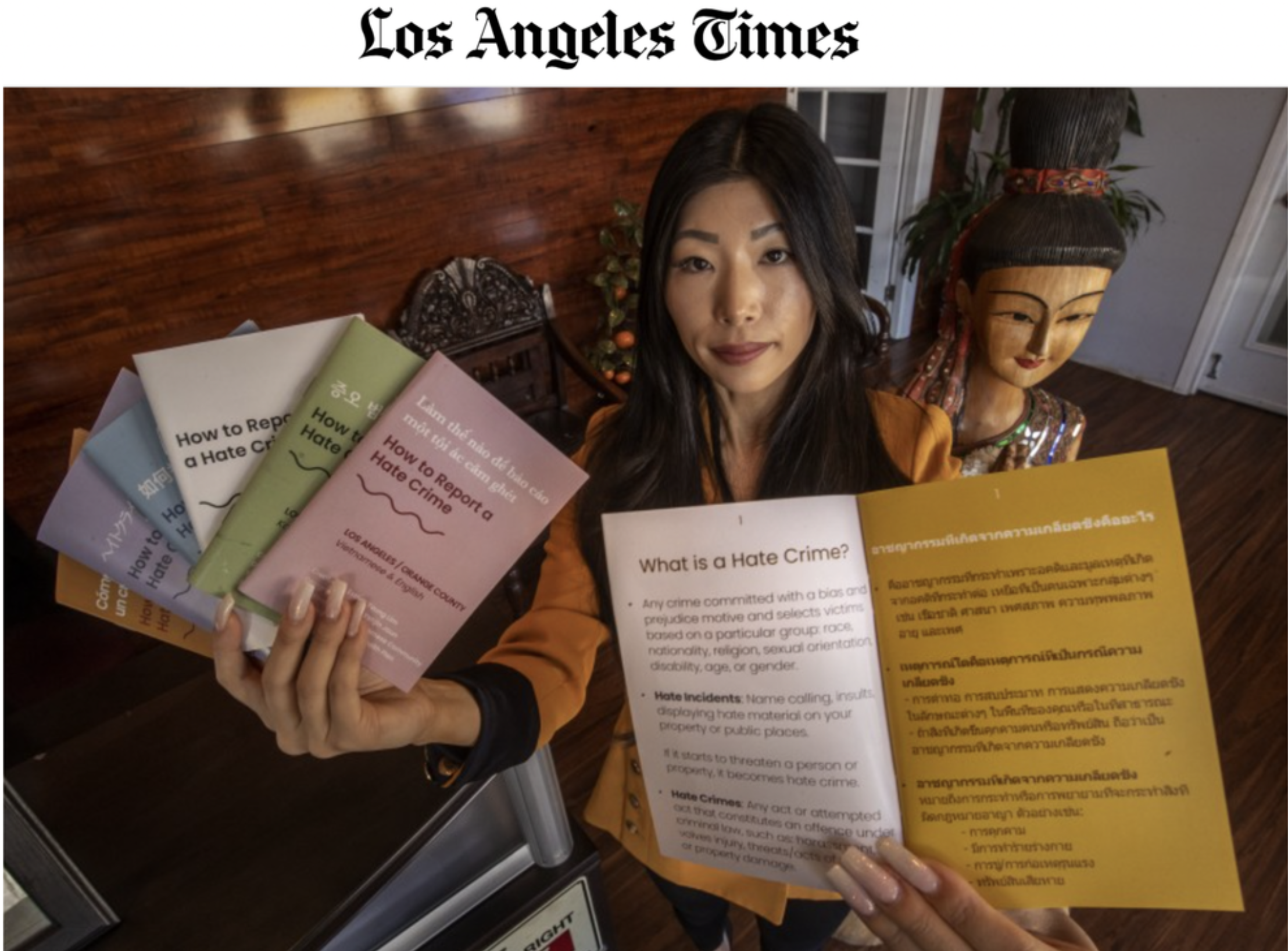Esther holding books and a pamphlet about hate crime featured in the LA Times, standing next to a wooden statue of a woman with traditional attire, in an indoor setting with plants and a wooden wall in the background.