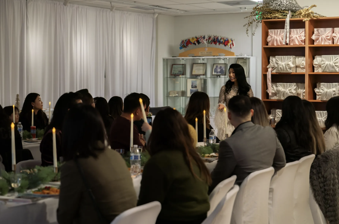 Esther standing and speaking in front of a seated audience about Hate Crime Booklets with tables decorated with candles and greenery featured in the San Gabriel Tribune.