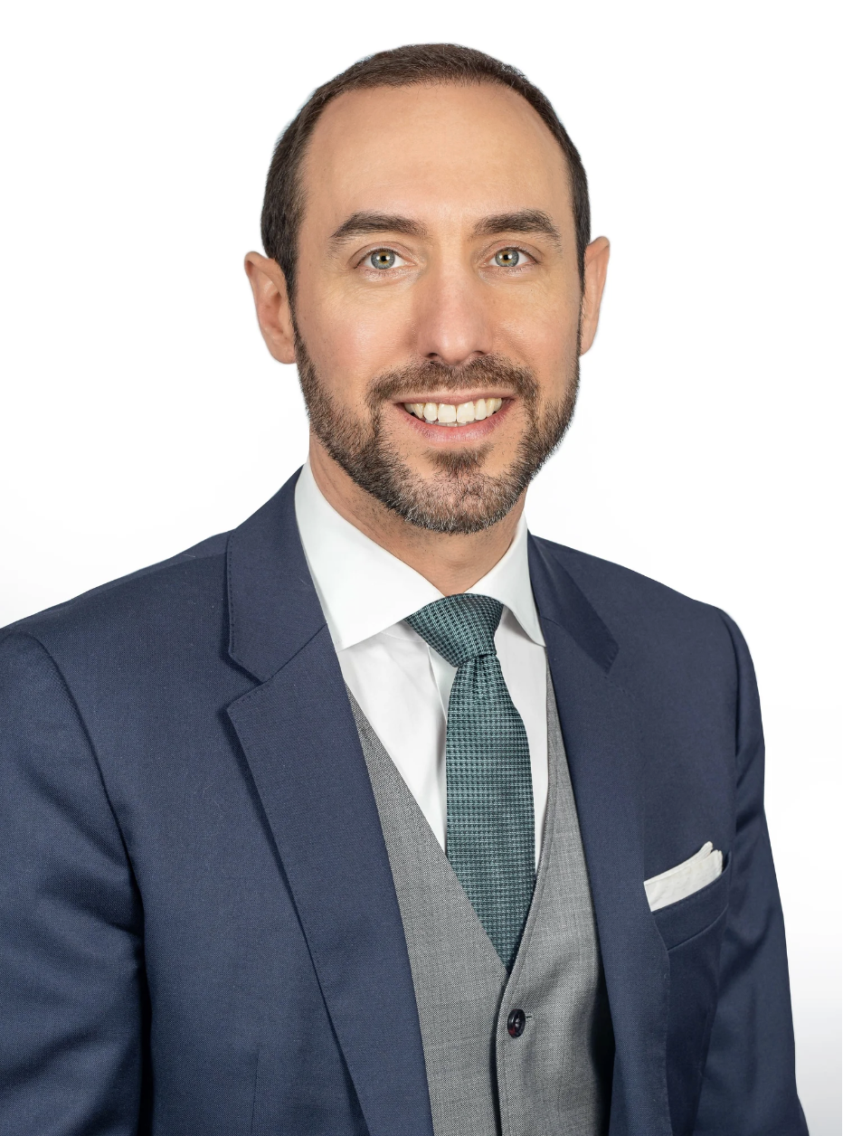 A man in a navy blue suit, white shirt, gray vest, and teal tie, smiling against a white background.