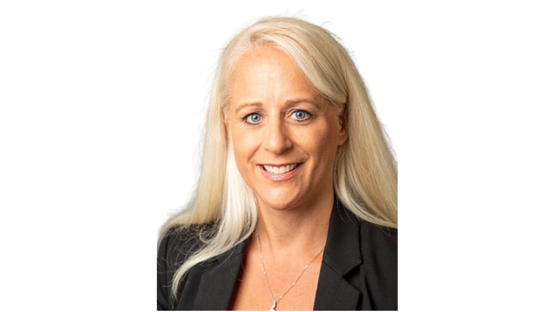Portrait of a smiling blonde woman with blue eyes wearing a black blazer and necklaces, against a white background.