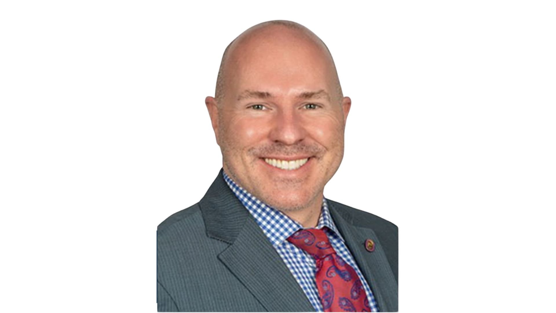 Portrait of a bald man with light skin, smiling, wearing a dark suit jacket, blue checked shirt, and a red patterned tie, against a white background.