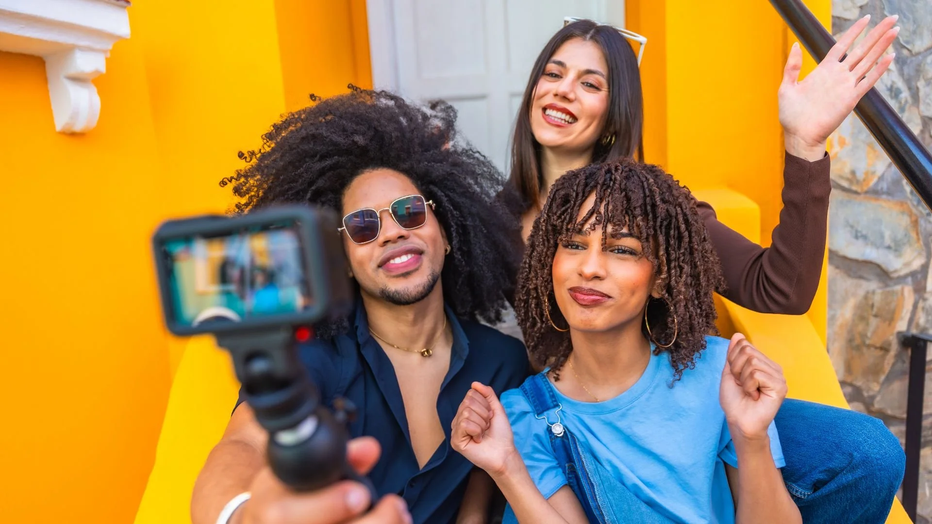 Three friends smiling and waving while filming a video blog with an action camera on a selfie stick against a yellow background - Encore Nationwide