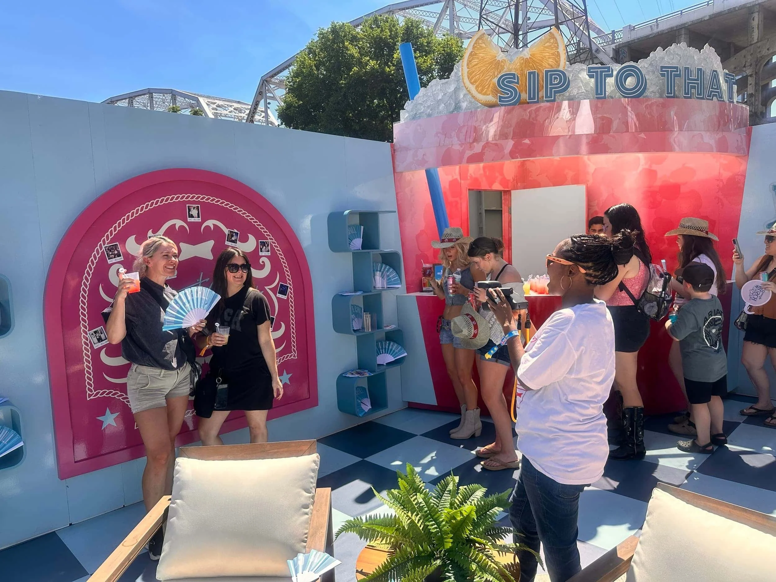 People holding drinks and fans while posing for a photo at a bright 'Sip To That' outdoor pop-up booth.