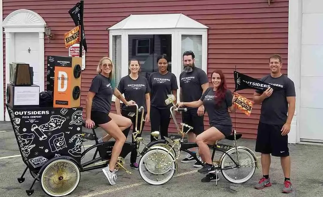 Outsiders Pizza Company promo team posing with custom brand bicycles and a pizza cart outside