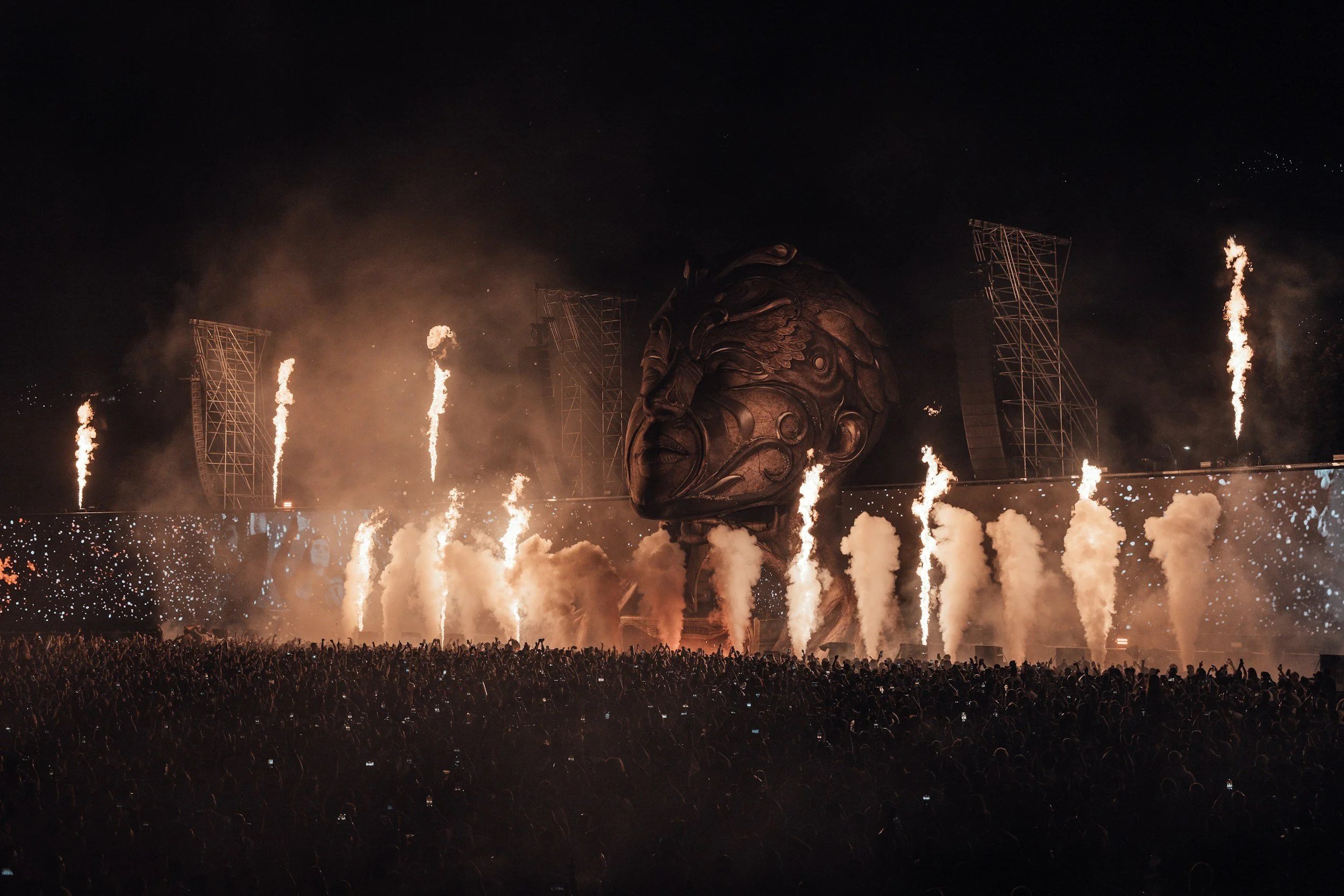Large crowd of concertgoers at a nighttime music festival featuring stage pyrotechnics, smoke effects, and a giant sculptural head design - Encore Nationwide